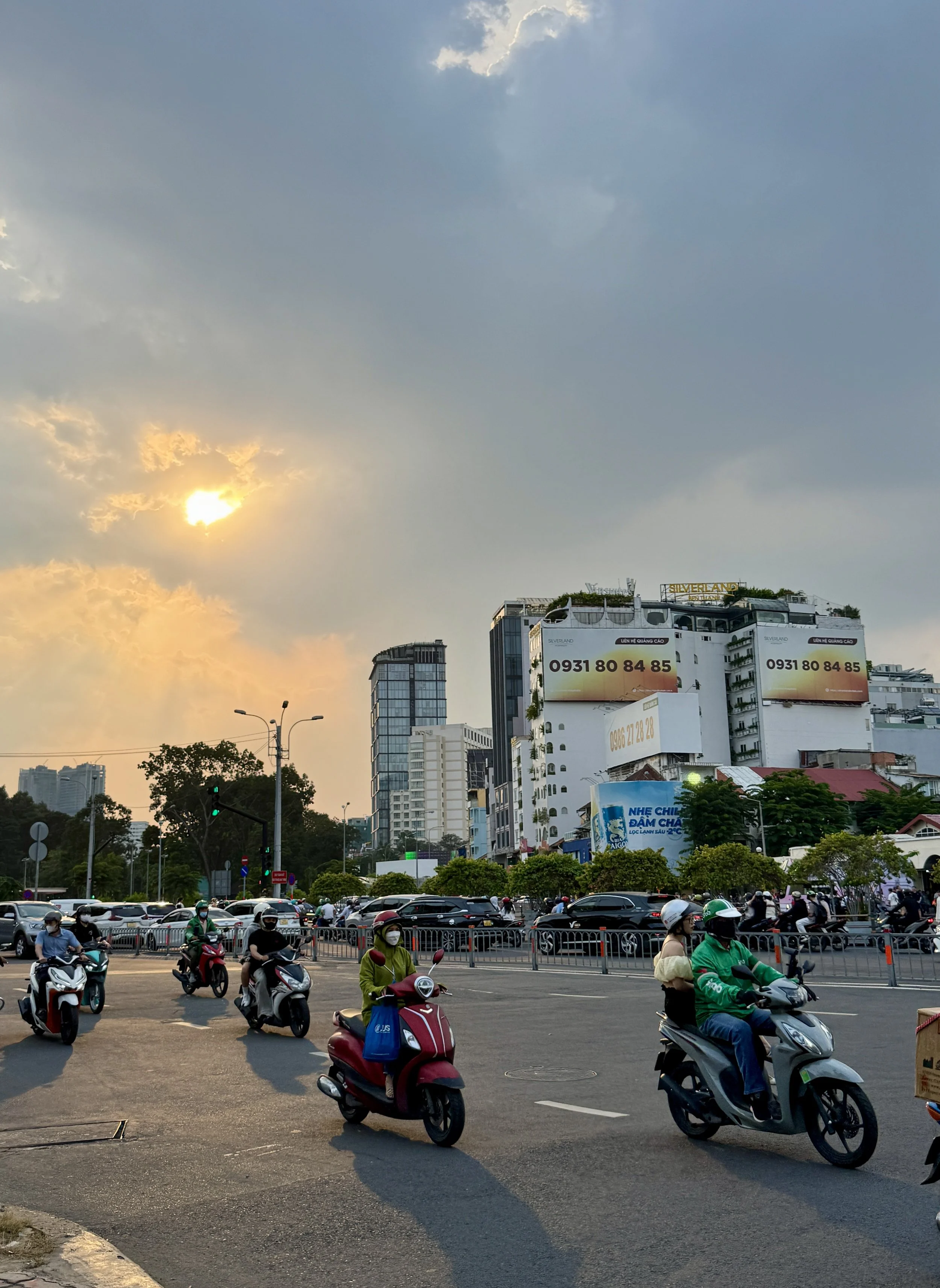Urban scene with people on motorbikes at an intersection, buildings in the background, and a cloudy sky with the sun partially obscured.