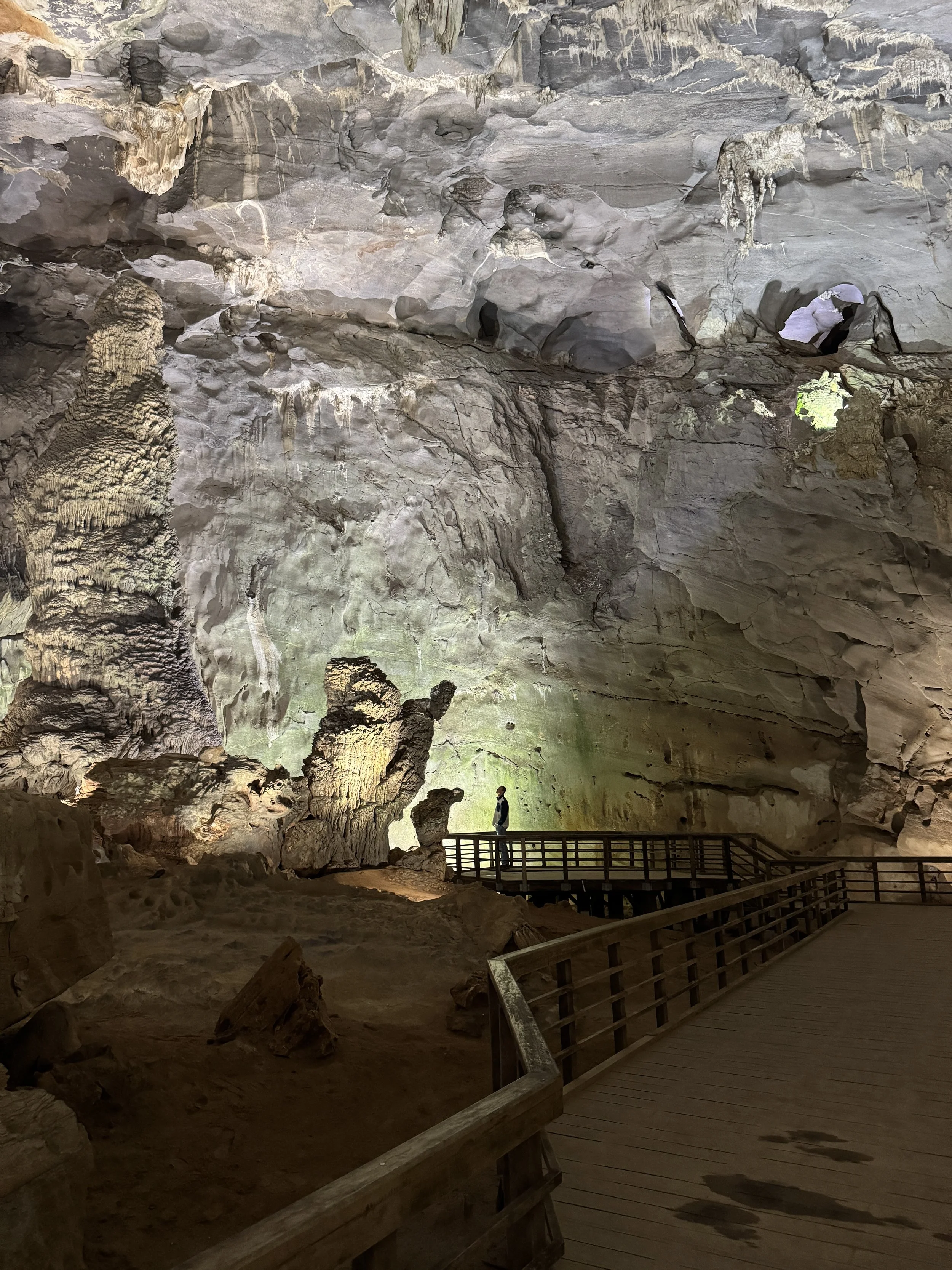 Person standing on a wooden walkway inside a large cave with rock formations and stalactites.