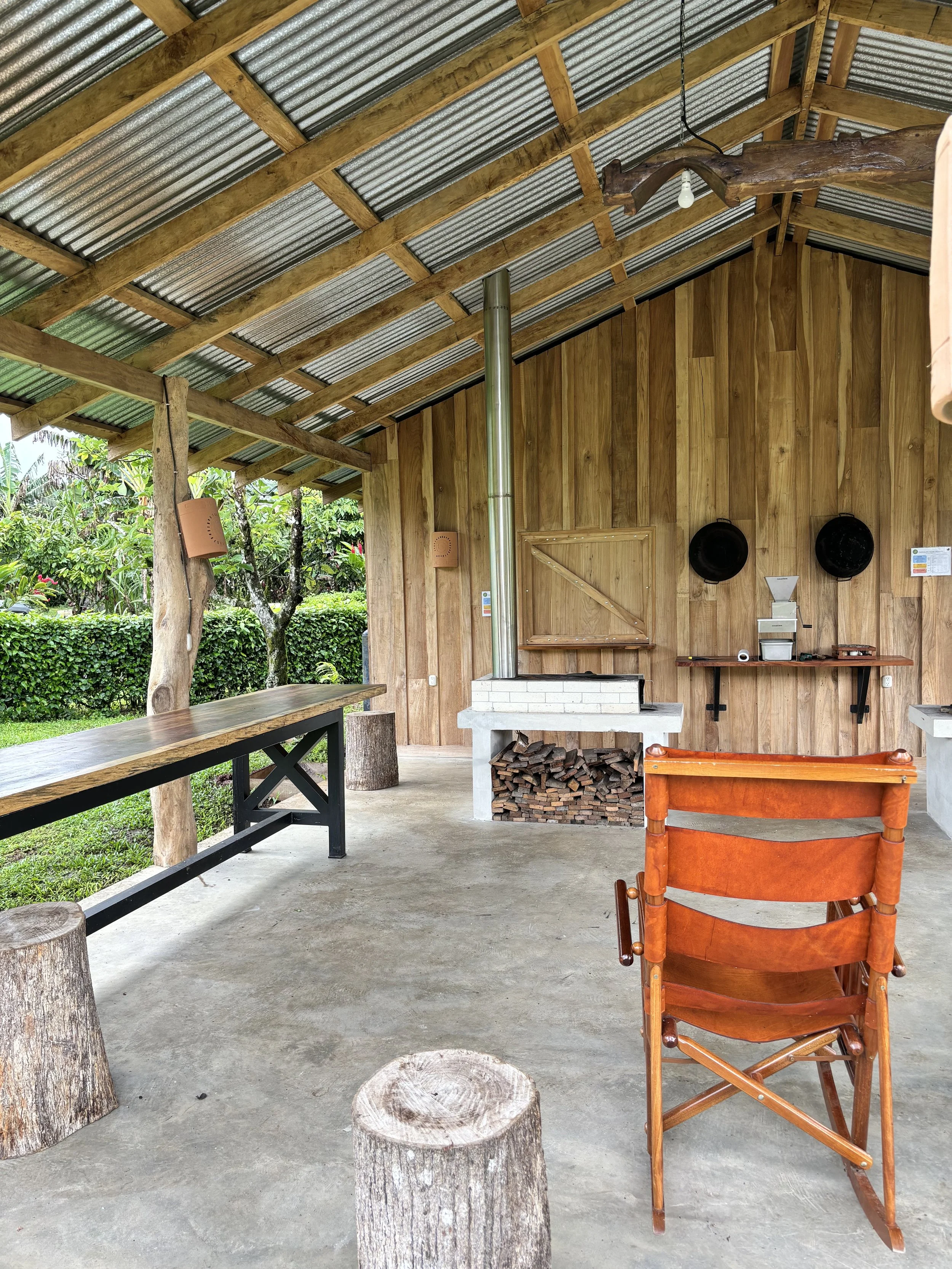 Rustic outdoor seating area with wooden walls, a corrugated metal roof, wooden benches, log stools, and a leather chair. There's a stacked wood pile and a chimney next to a wall-mounted shelf with various items.