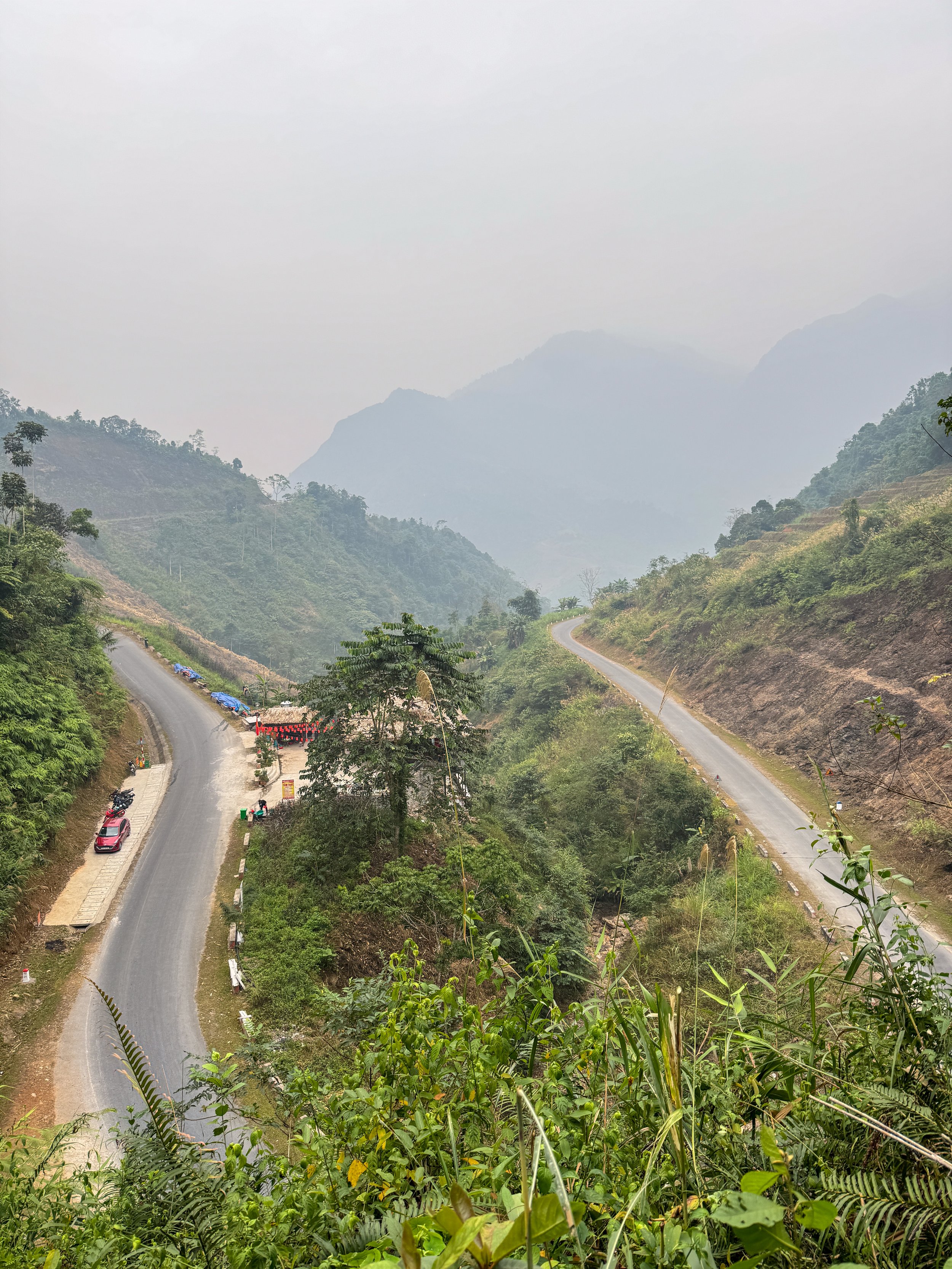 Mountain road winding through lush green hills, with misty mountains in the background. A pink car parked beside the road, surrounded by vegetation.
