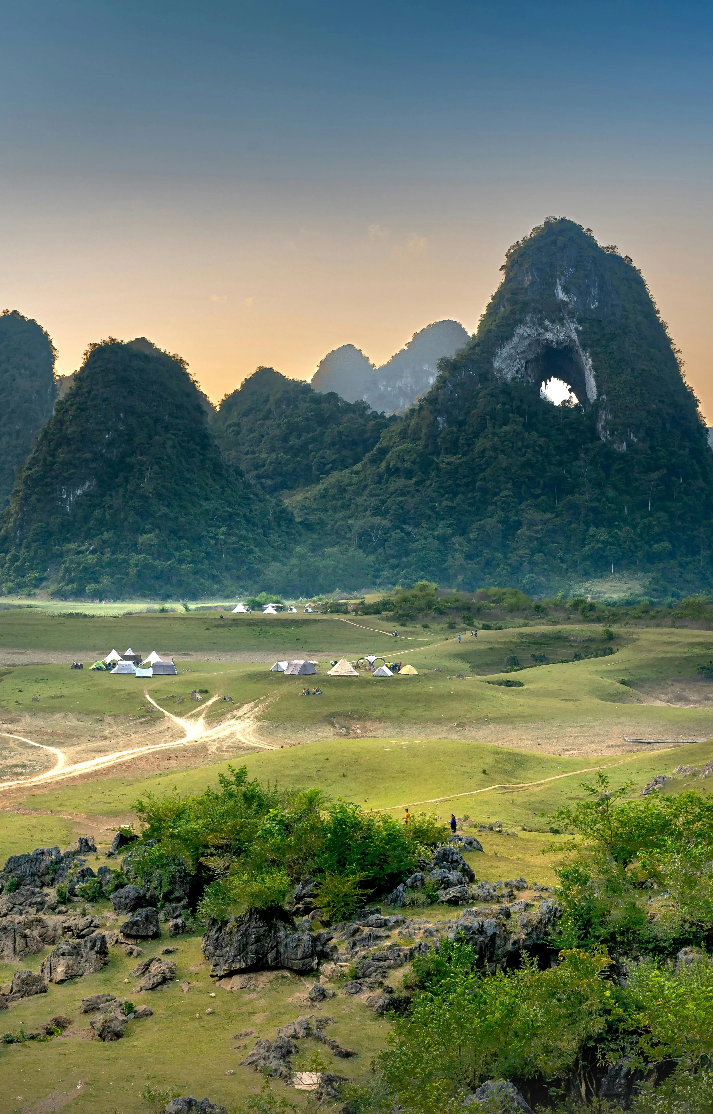 Scenic landscape with green hills, rocky formations, tents, and limestone mountains in the background.