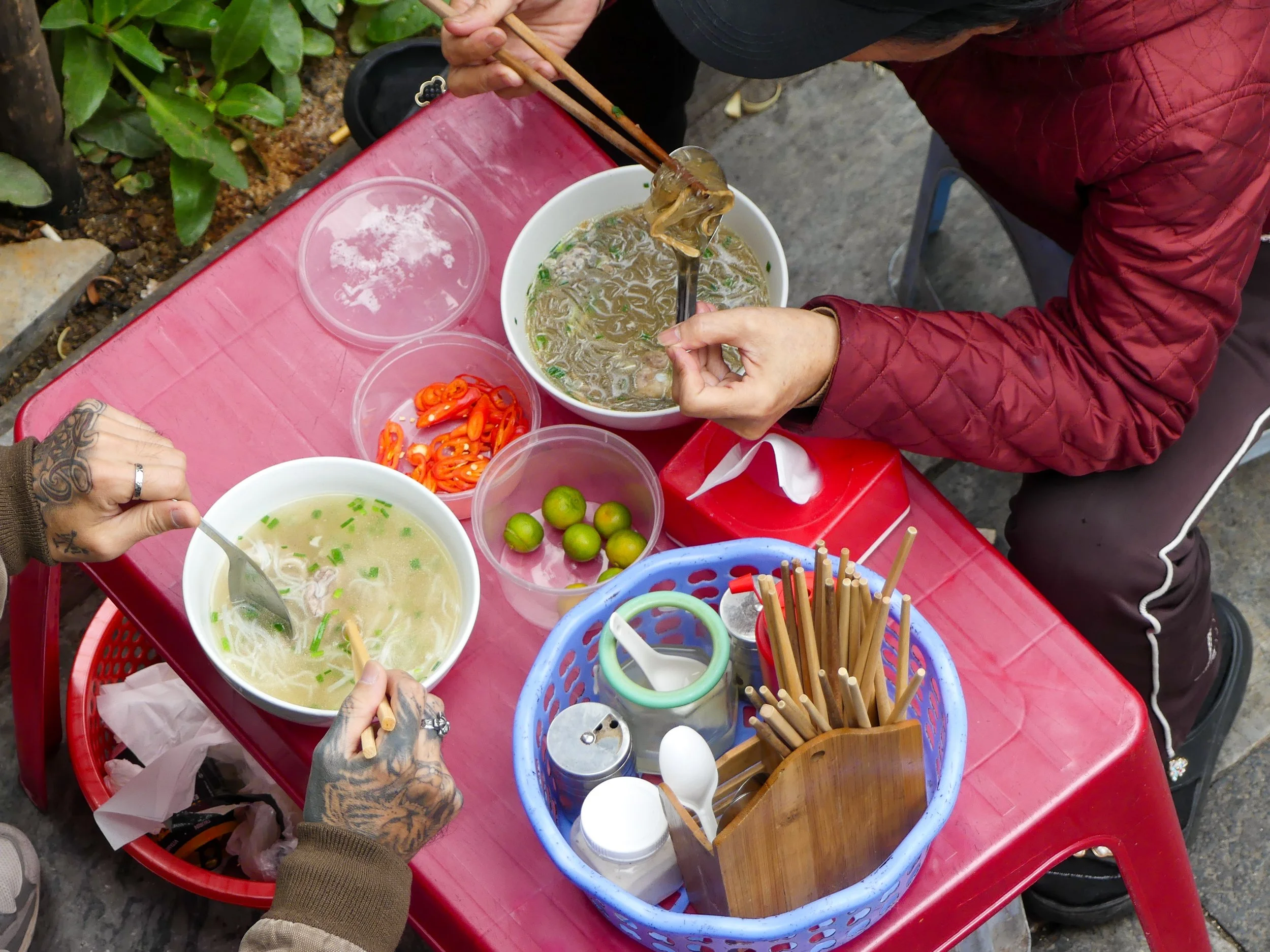 Two people eating noodle soup with chopsticks and spoons sitting at a red plastic table, with small bowls of sliced chili peppers and limes, and a basket with chopsticks and utensils beside them.