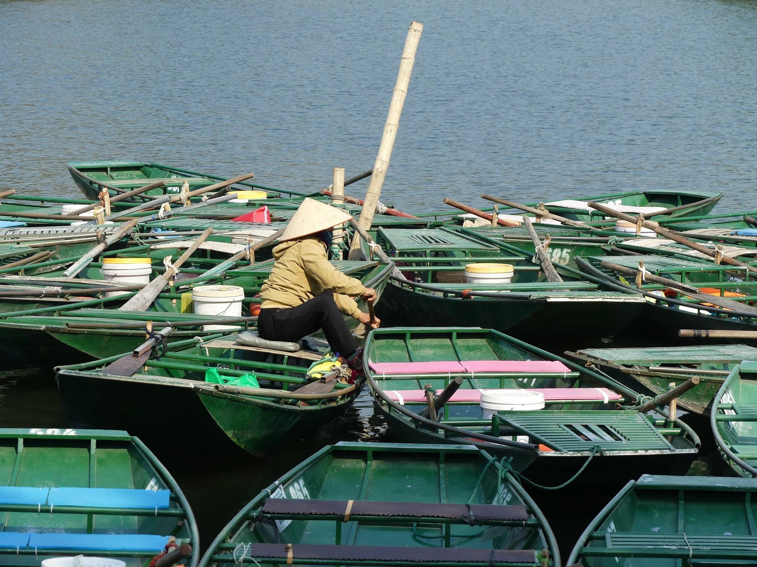 Person wearing a straw hat sitting among green wooden boats on water.