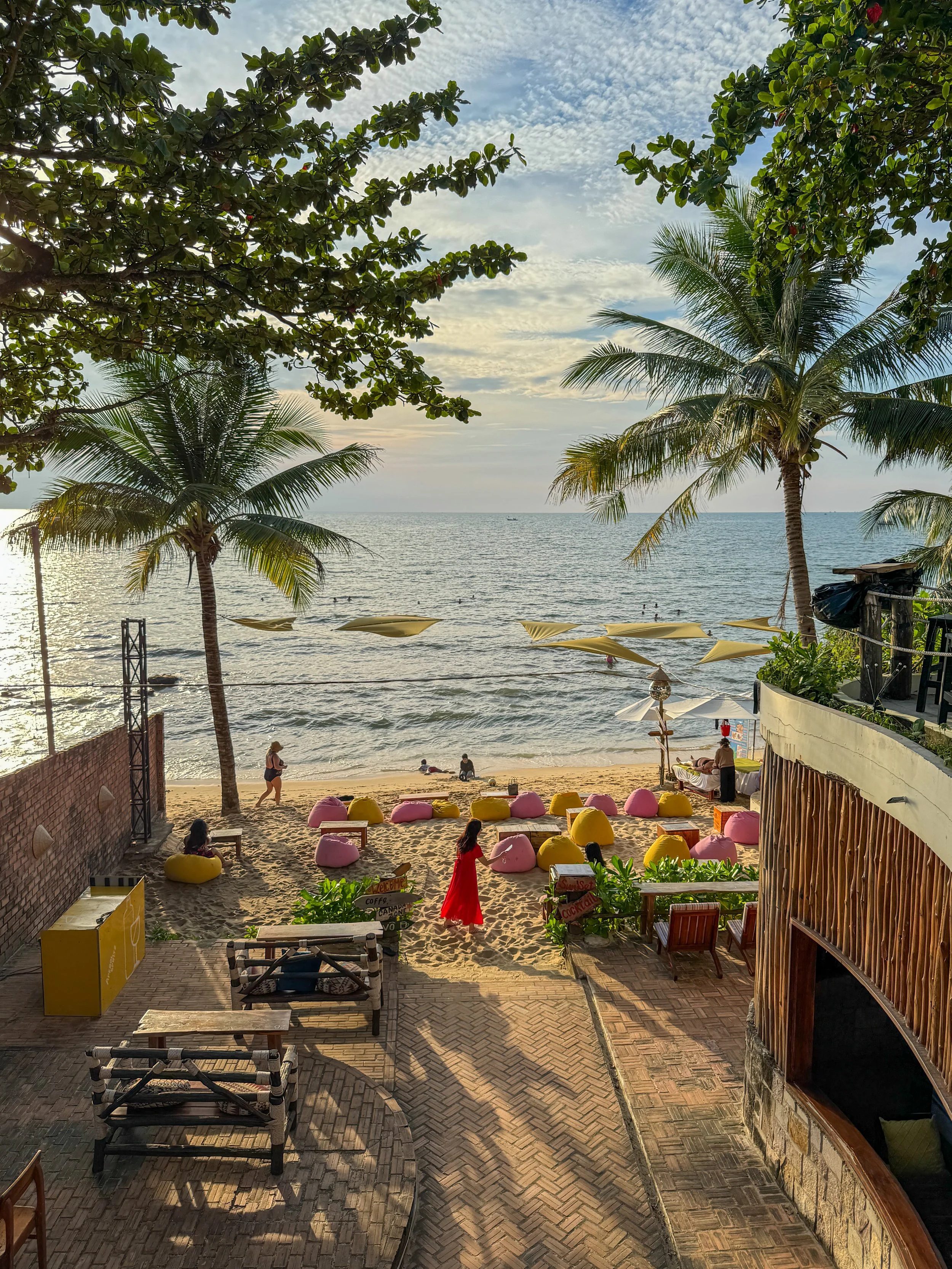 Beach scene with palm trees, bean bag chairs, a person in a red dress, and ocean view on a sunny day.