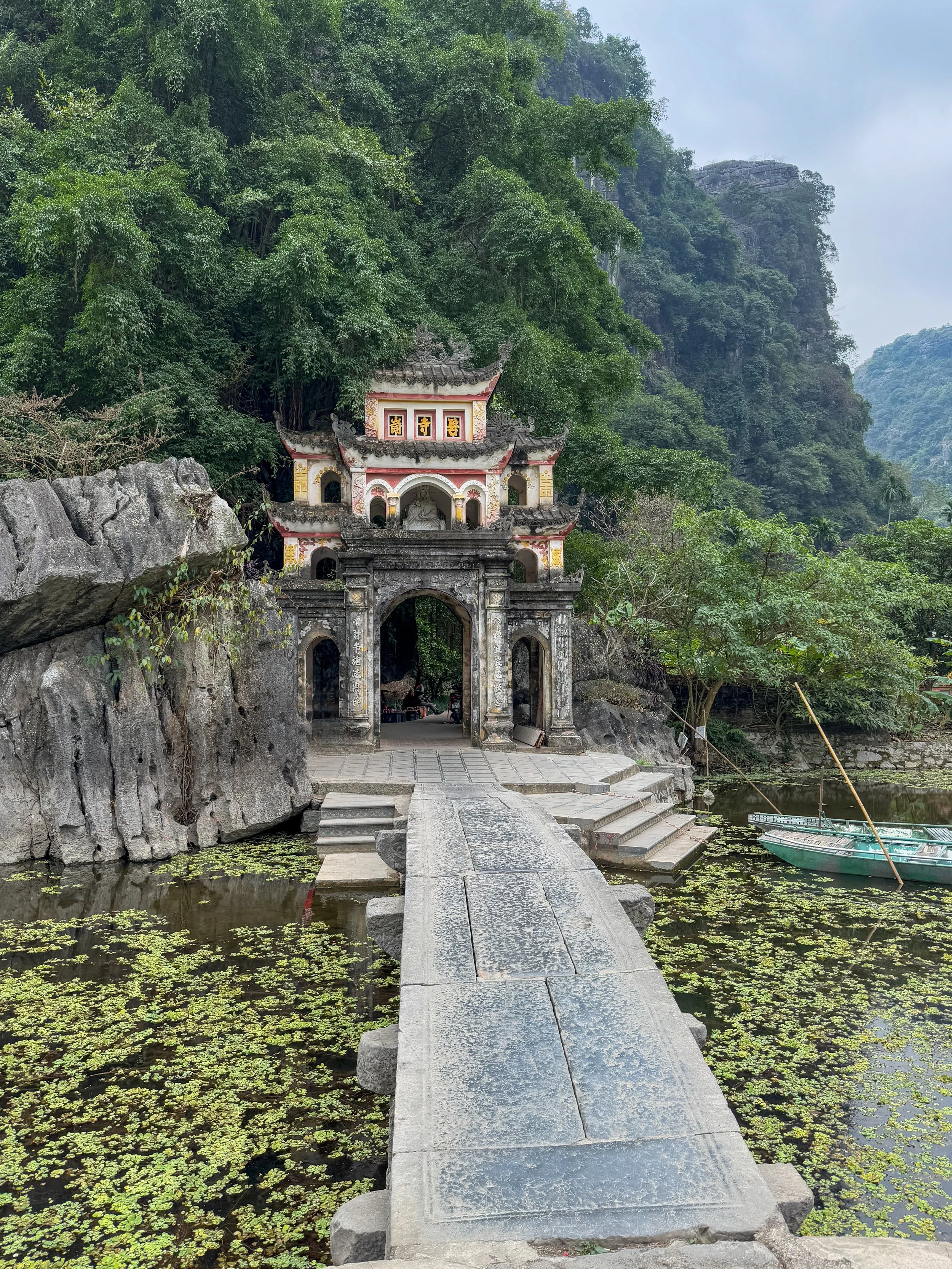 Scenic view of an ancient stone gate with arched openings and decorative elements, set amidst lush greenery. A stone walkway leads to the gate over a pond with floating vegetation. A small boat with oars is moored to the side.