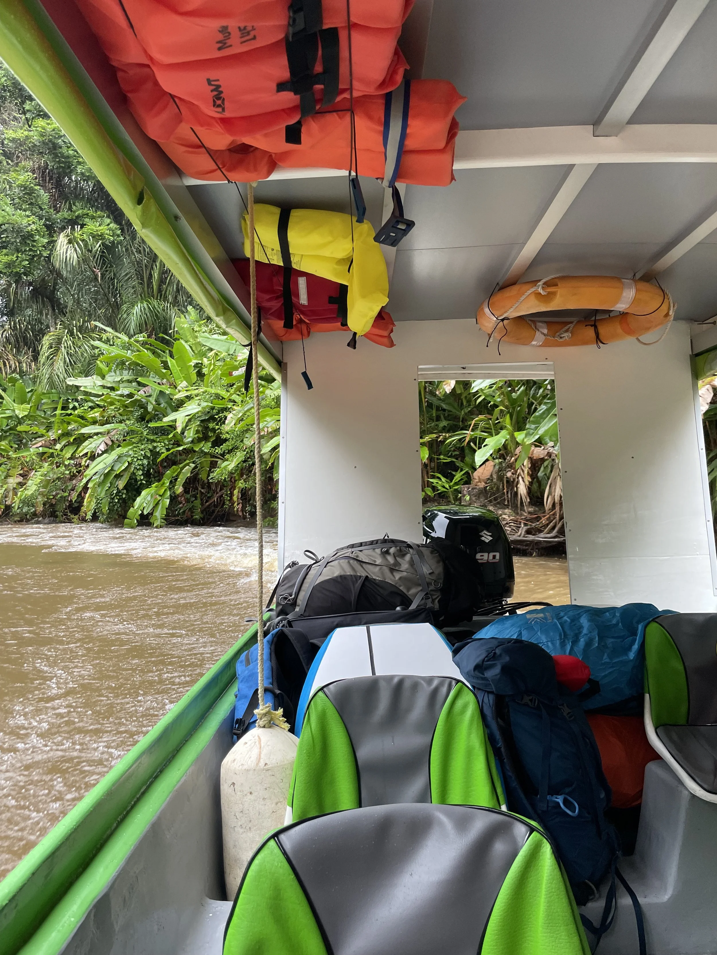 Interior view of a small boat with life jackets on the ceiling, backpacks on the seats, and lush greenery outside.