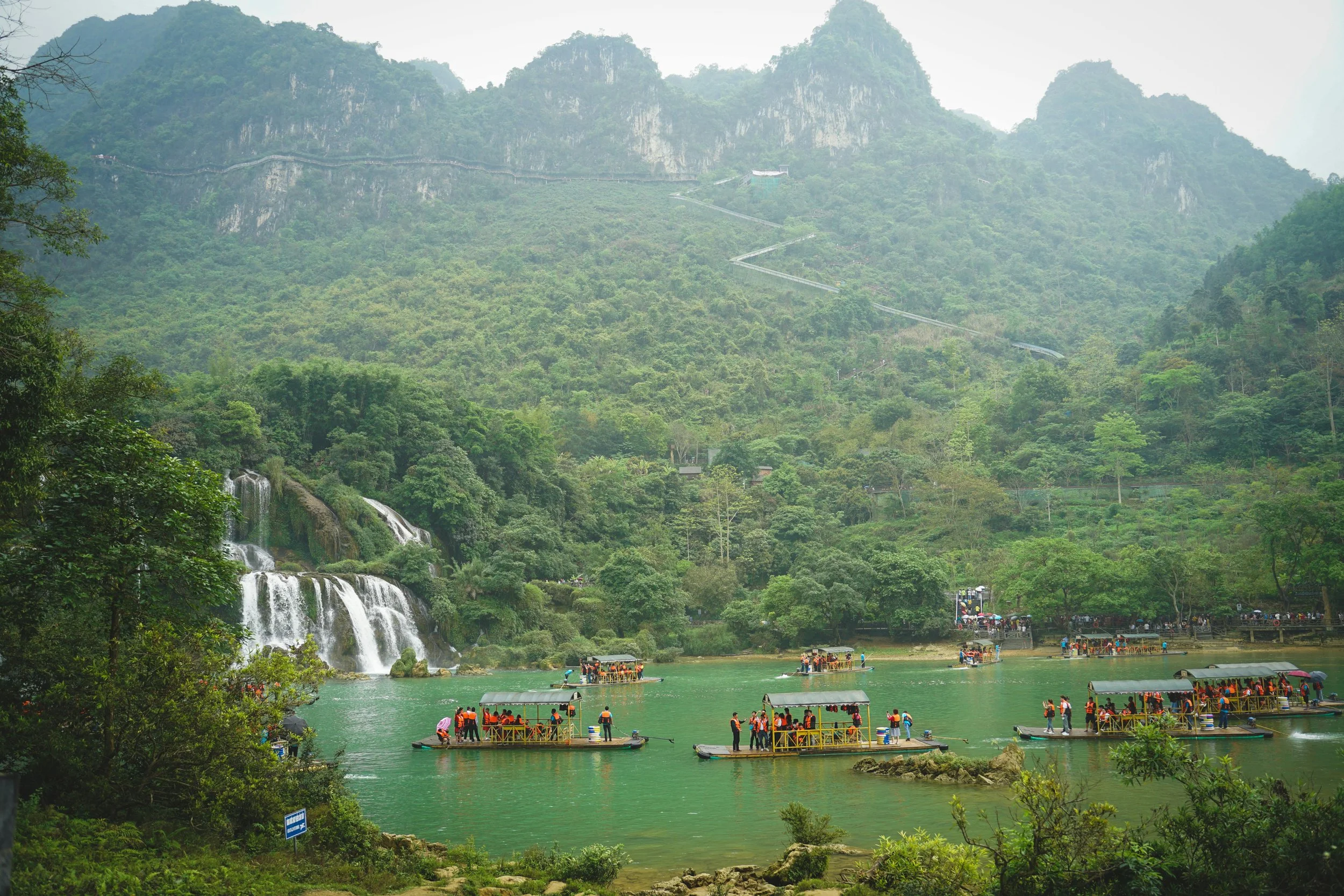 Waterfall with multiple tiers in a green mountainous landscape, surrounded by trees, with several boats carrying tourists on a river in the foreground.