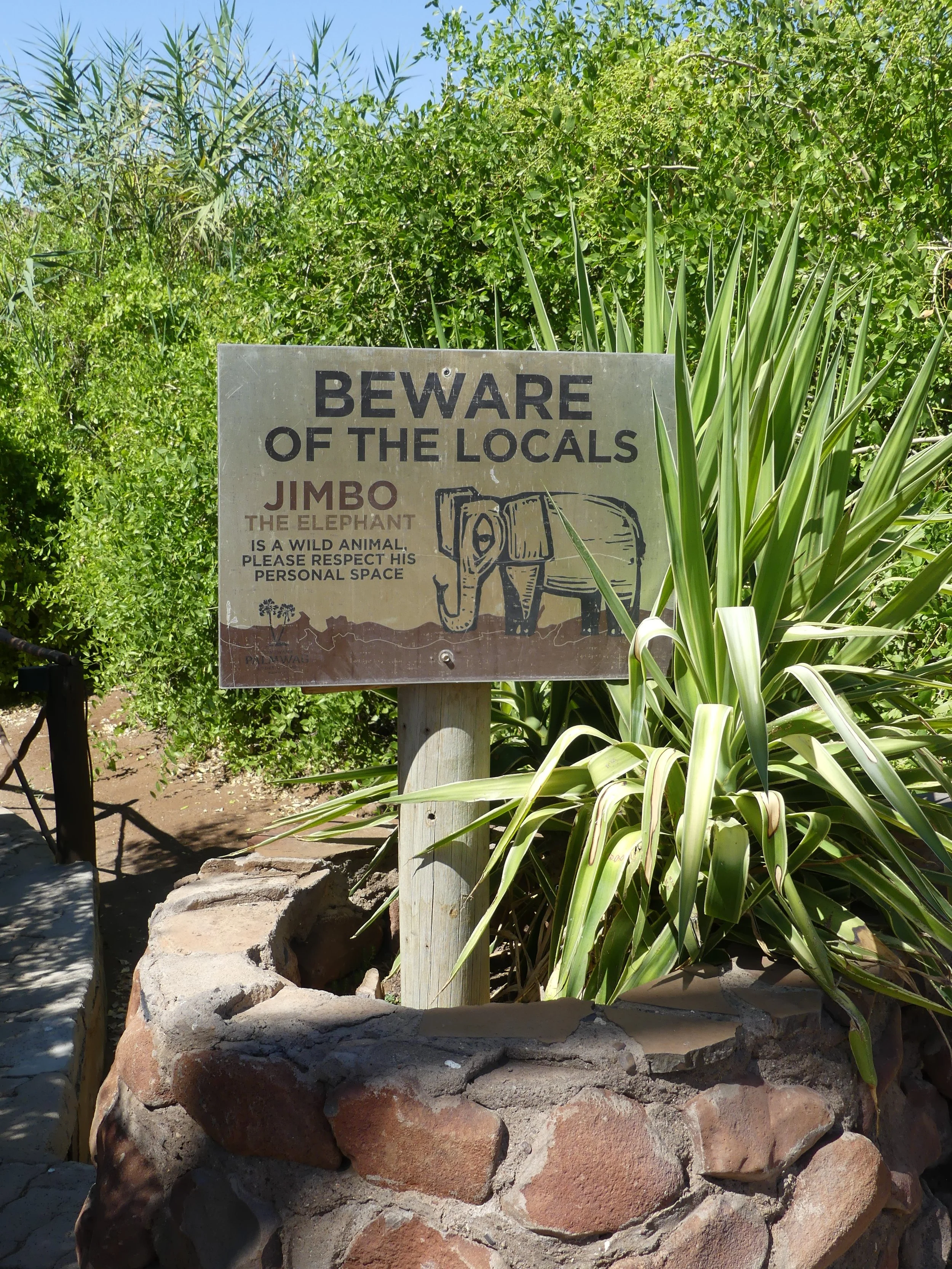 A warning sign in a natural setting warns visitors to beware of the local wildlife, particularly an elephant named Jimbo. A visit of the rare desert elephant to a campsite in Palmwag Namibia