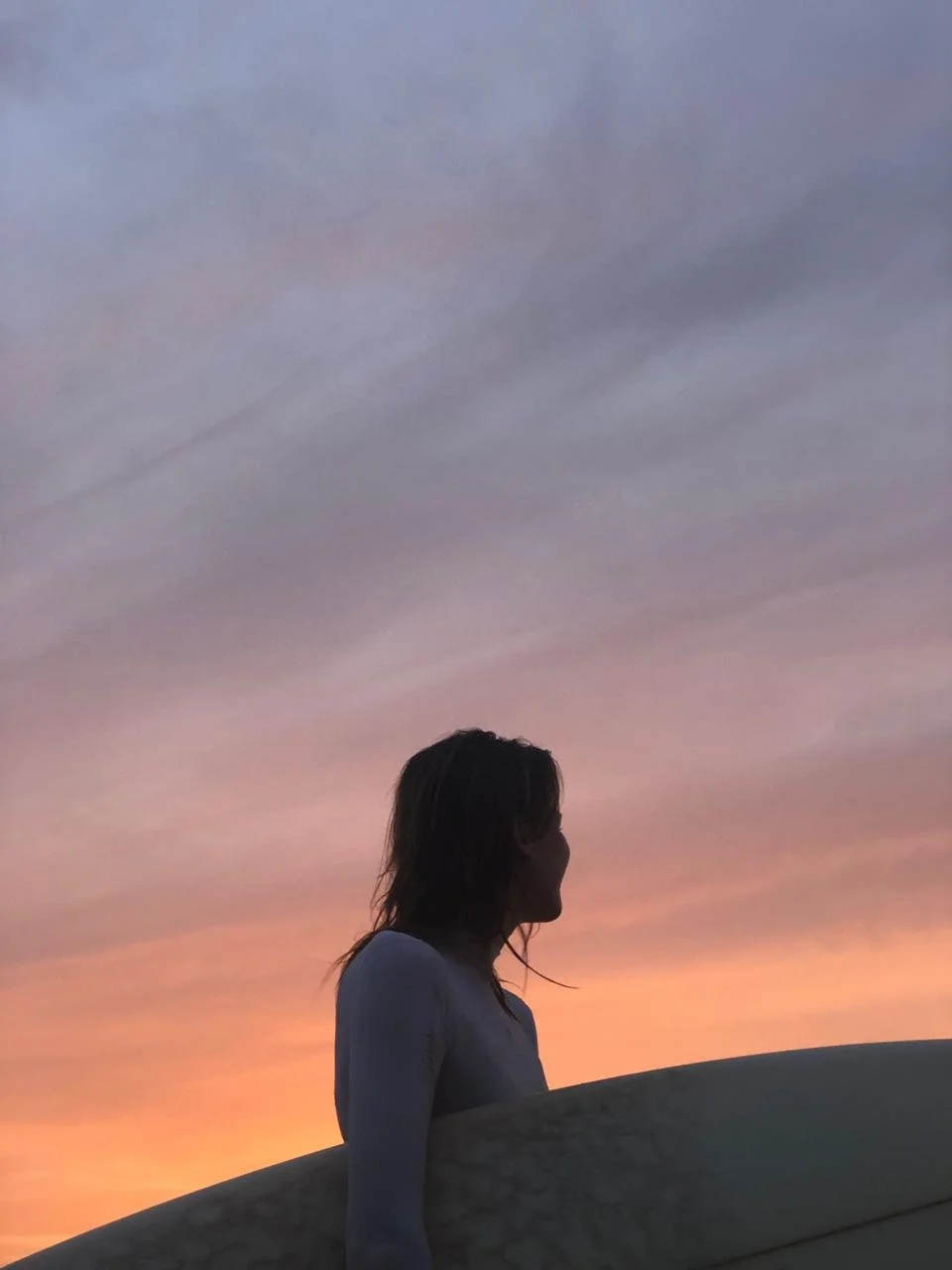 Silhouette of a person with long hair holding a surfboard against a colorful sunset sky.