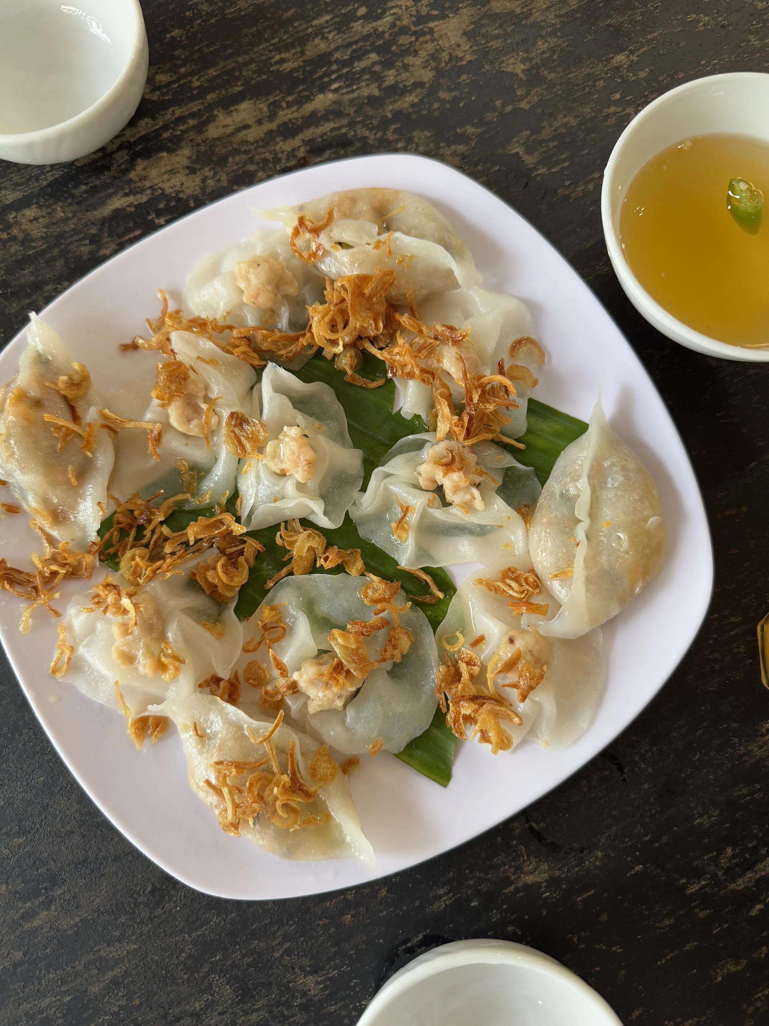 A plate of steamed dumplings topped with fried onions, served on a banana leaf, with a bowl of dipping sauce on the side.