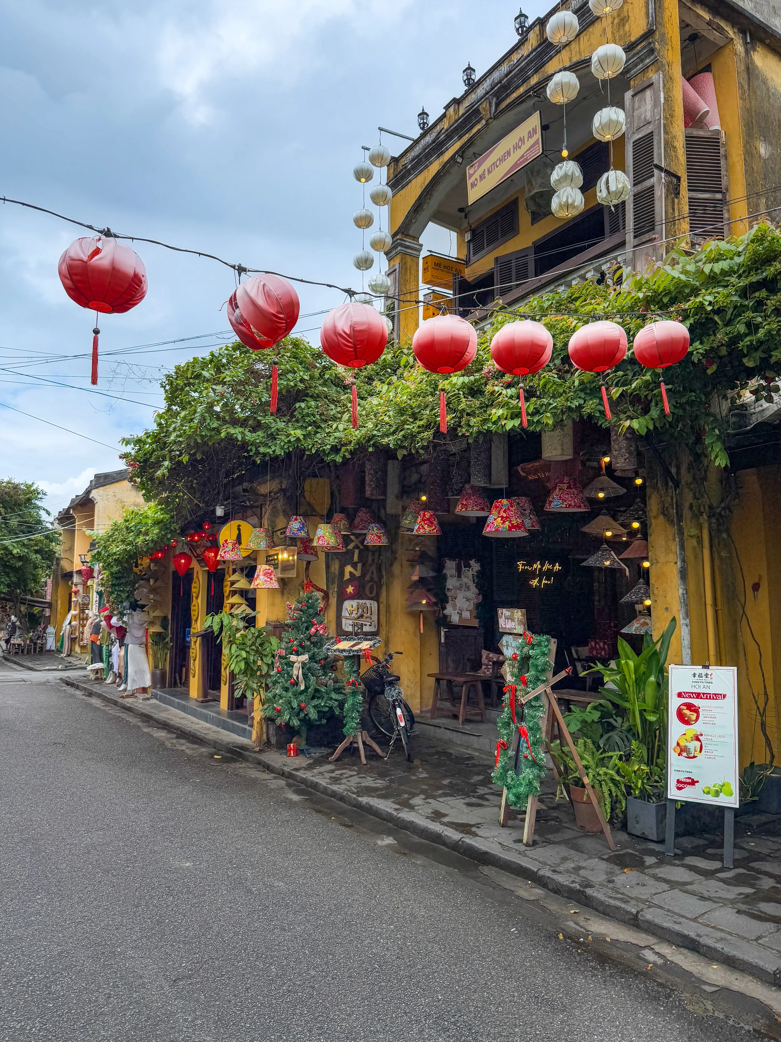 Street scene in Hoi An, Vietnam, featuring a yellow historic building adorned with red and white lanterns, potted plants, and festive decorations. The sidewalk displays various decorative items, and a motorbike is parked nearby under overcast skies.