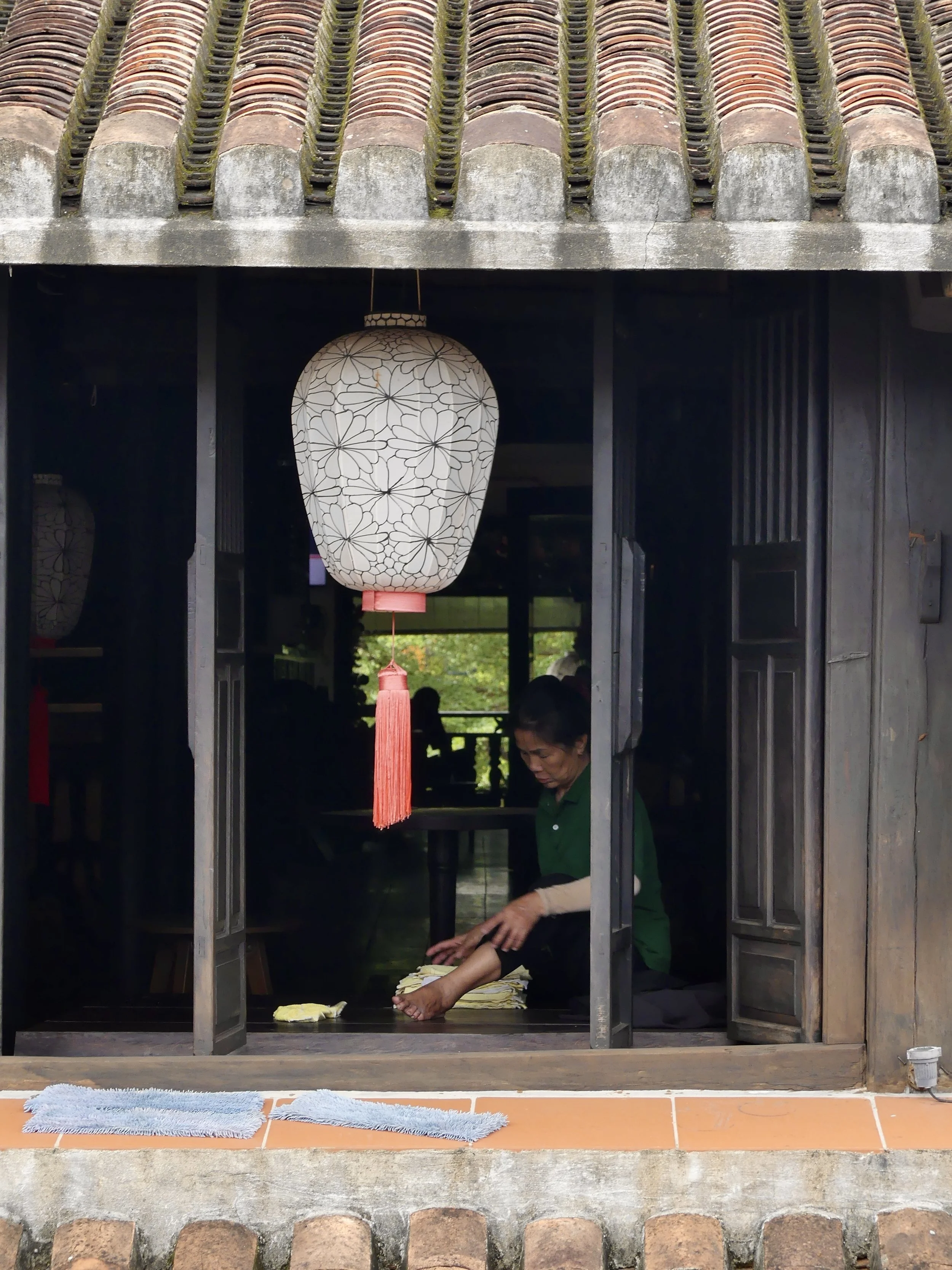 A person sitting inside a traditional wooden building with a tiled roof, folding fabric. A decorative paper lantern hangs at the entrance.