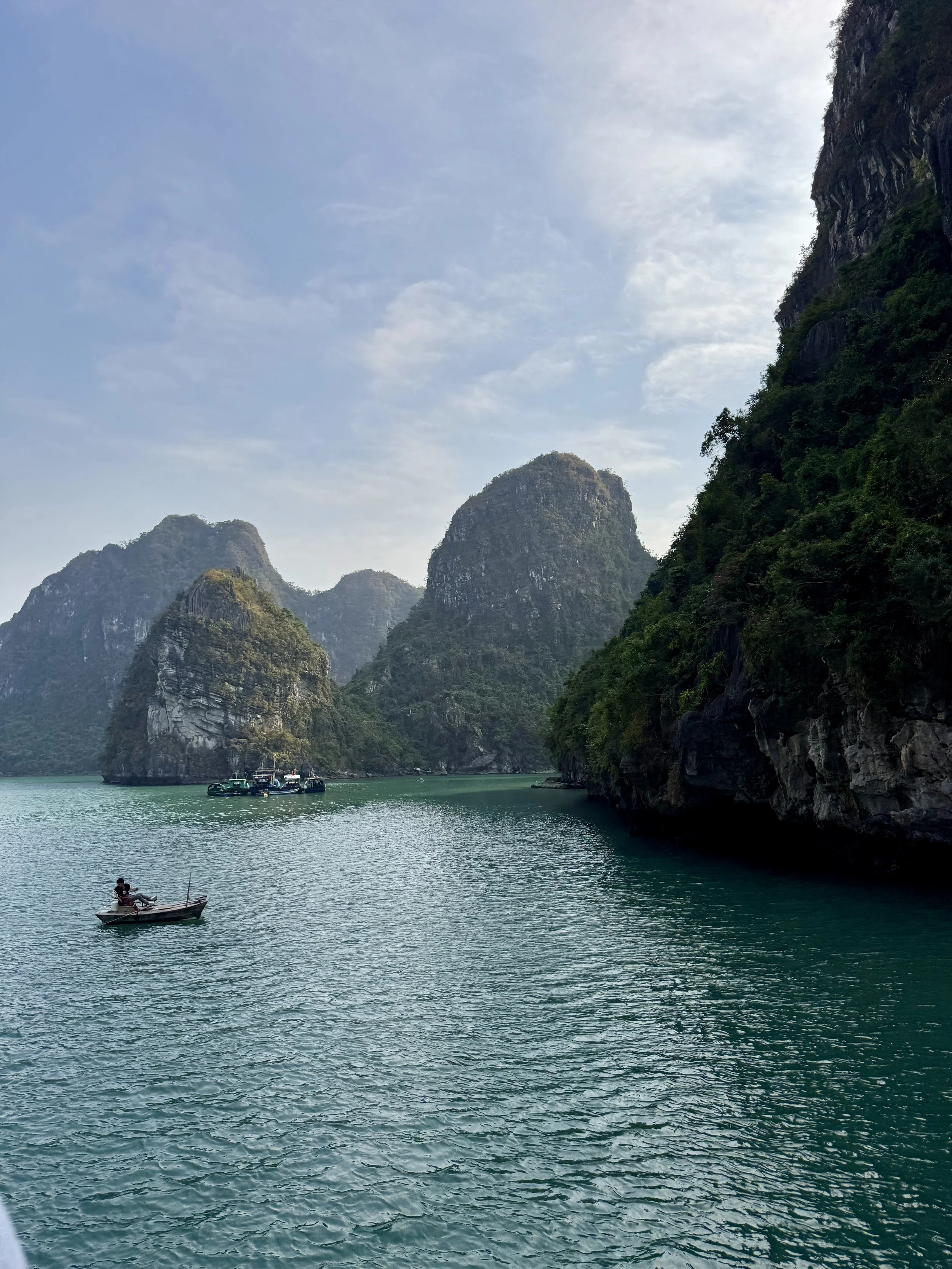 Scenic view of a bay with limestone karst formations and a person rowing a small boat on calm green waters under a partly cloudy sky.