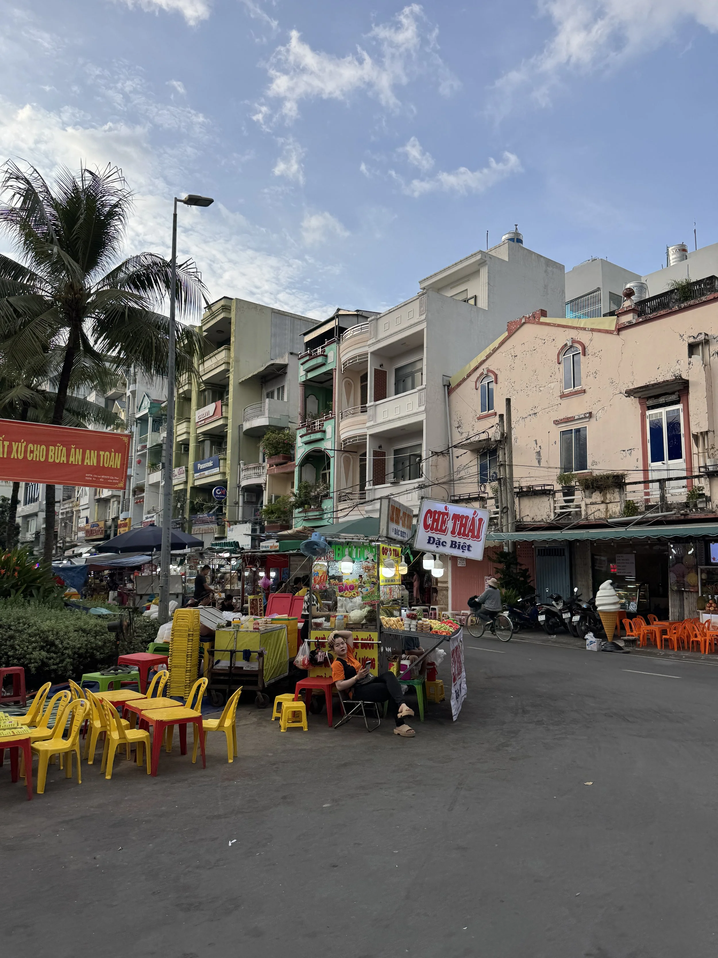 Outdoor street market in an urban setting with colorful plastic chairs and tables, food stalls, a palm tree, and multi-story buildings in the background.