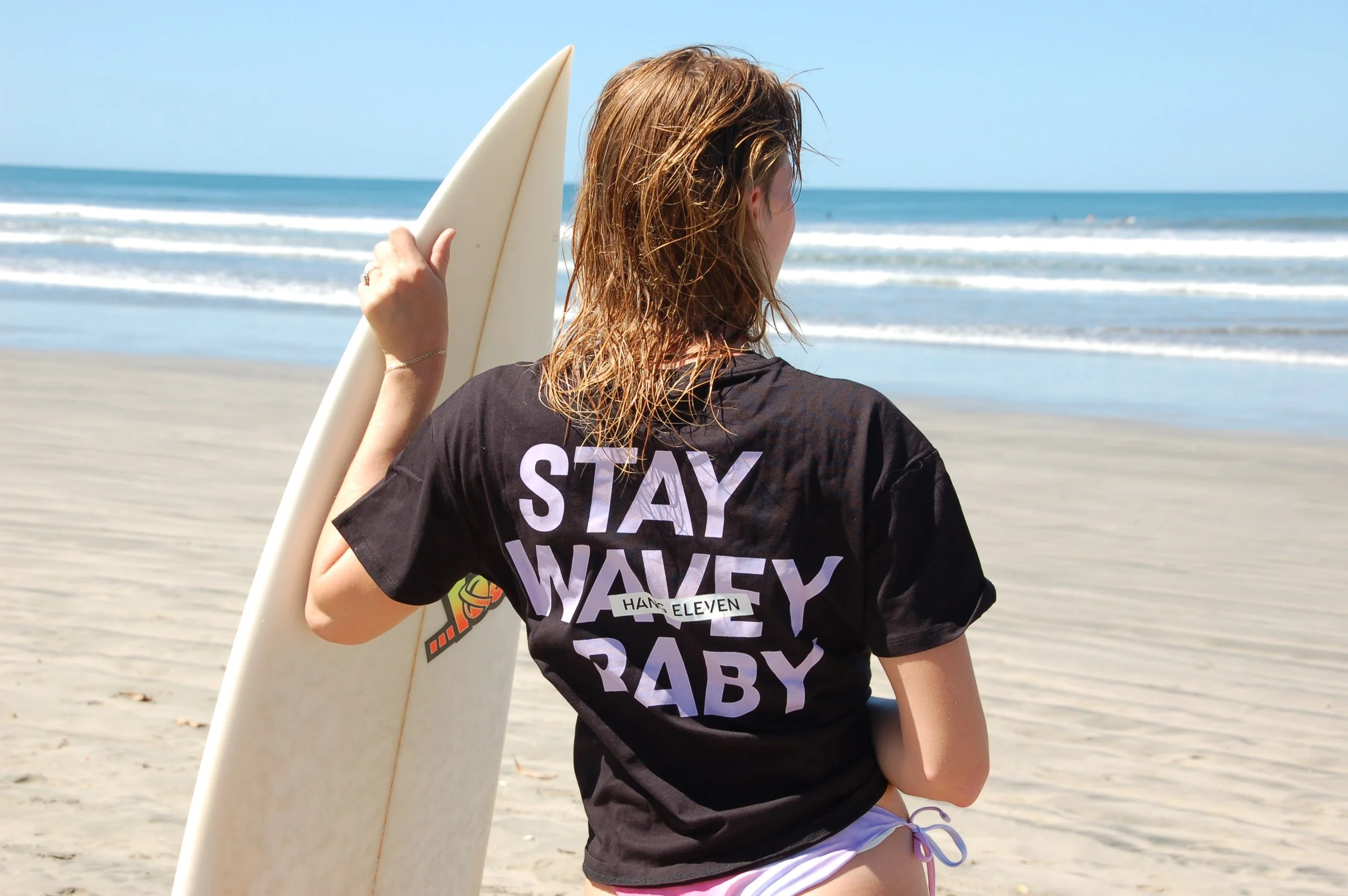 Person holding a surfboard on a beach, wearing a black shirt with "Stay Wavey Baby" printed on it.