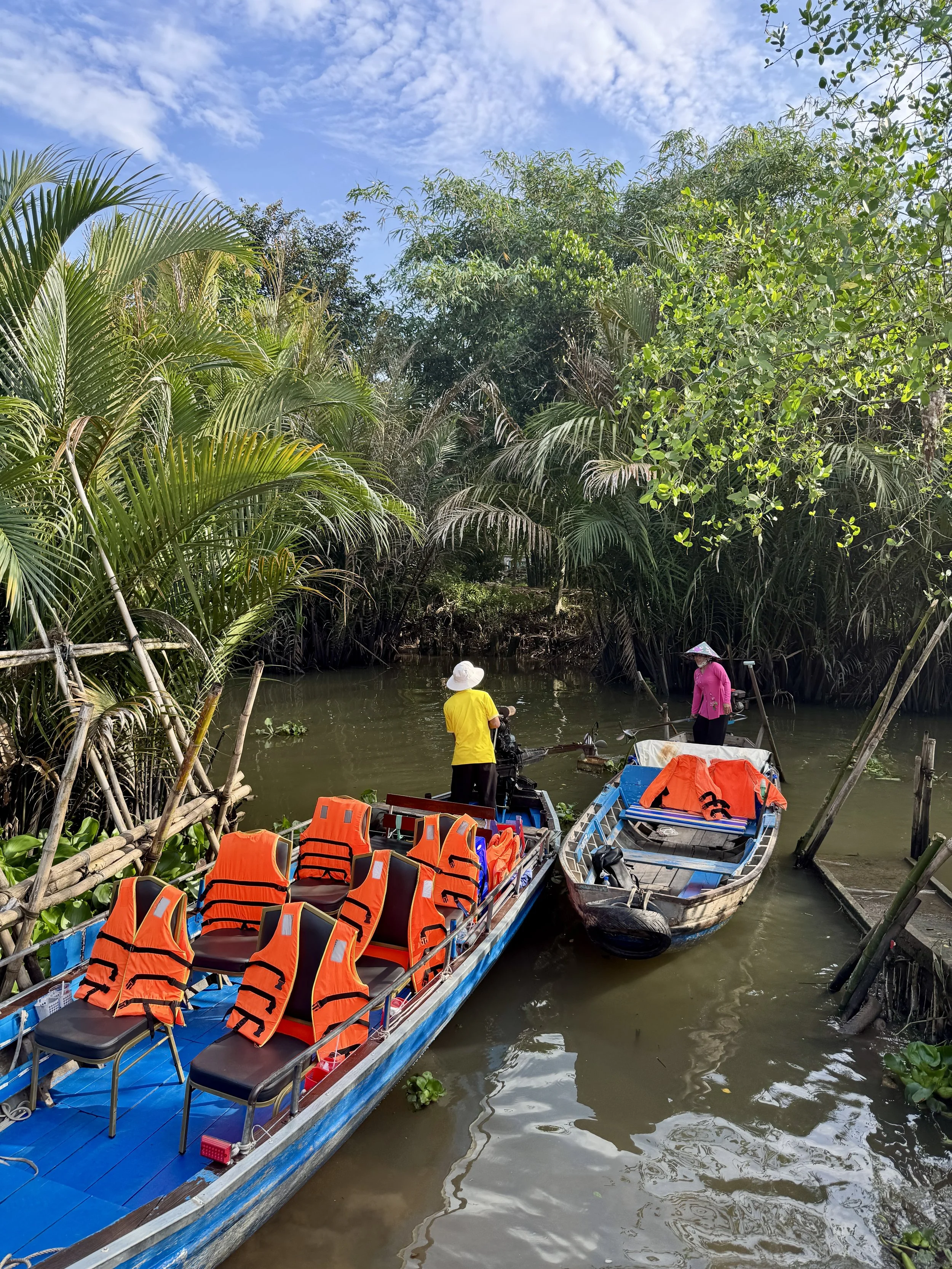 Two boats with orange life vests docked on a river, surrounded by lush green tropical foliage. Two people wearing hats stand on each boat, under a partially cloudy sky.