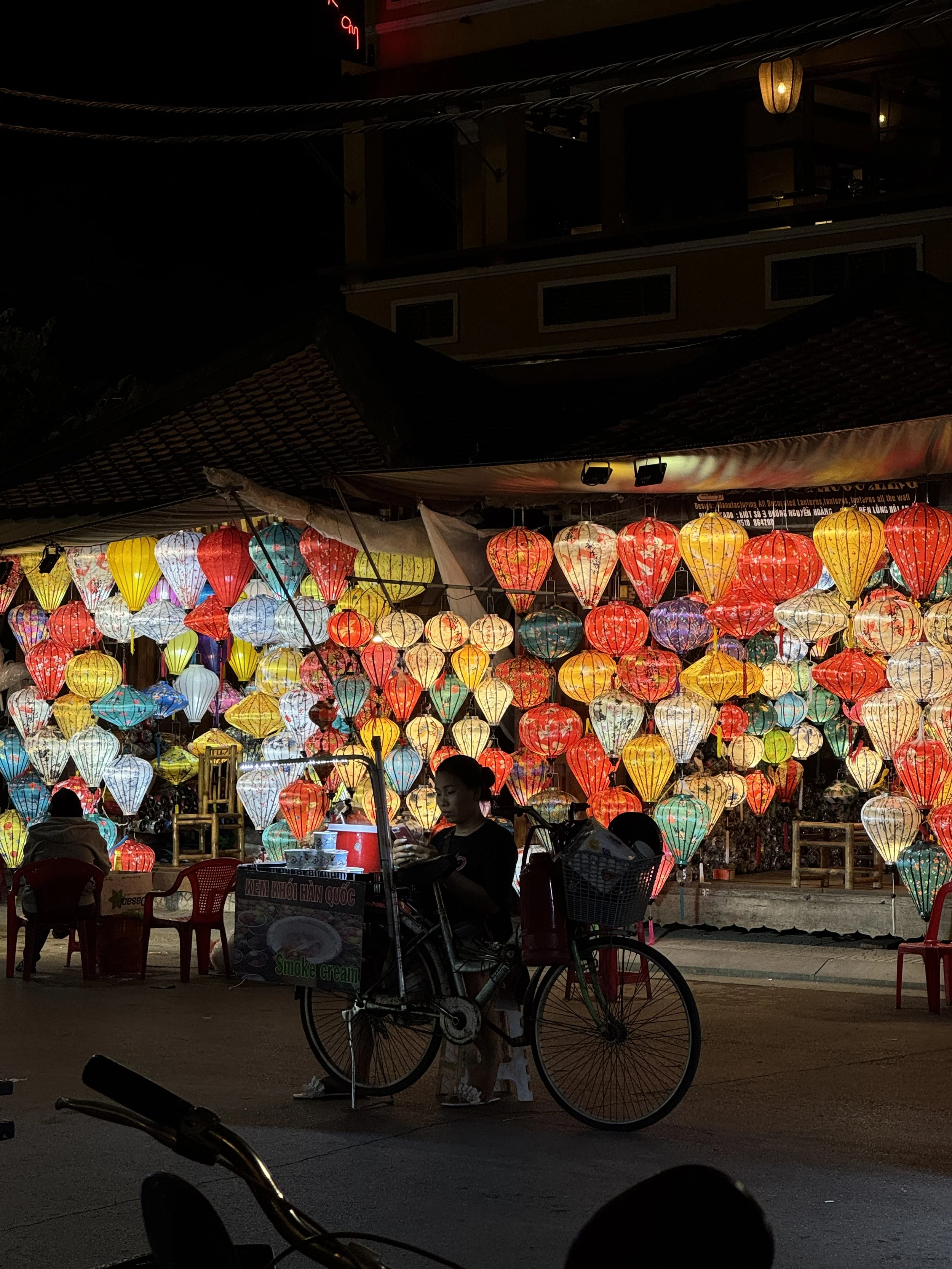 Colorful lanterns displayed at night with red chairs and a person sitting on a bicycle cart.