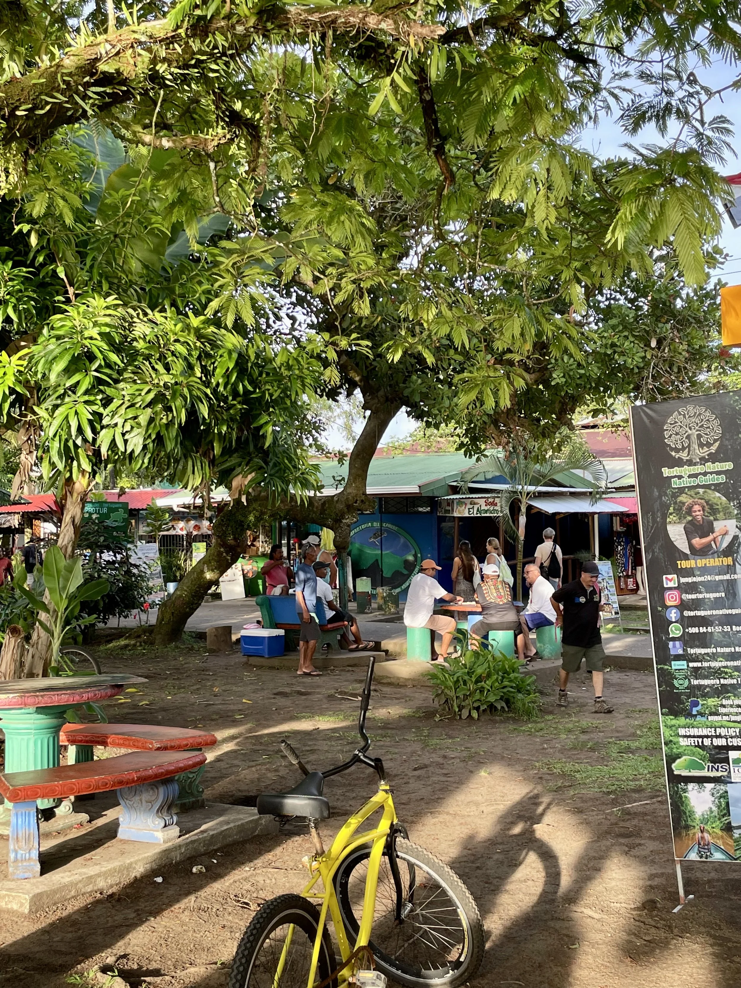 Park scene with a group of people sitting at tables under a large tree. There's a yellow bicycle in the foreground, colorful benches, and a banner promoting a tour operator with contact details. The area is surrounded by greenery and there are buildings in the background.