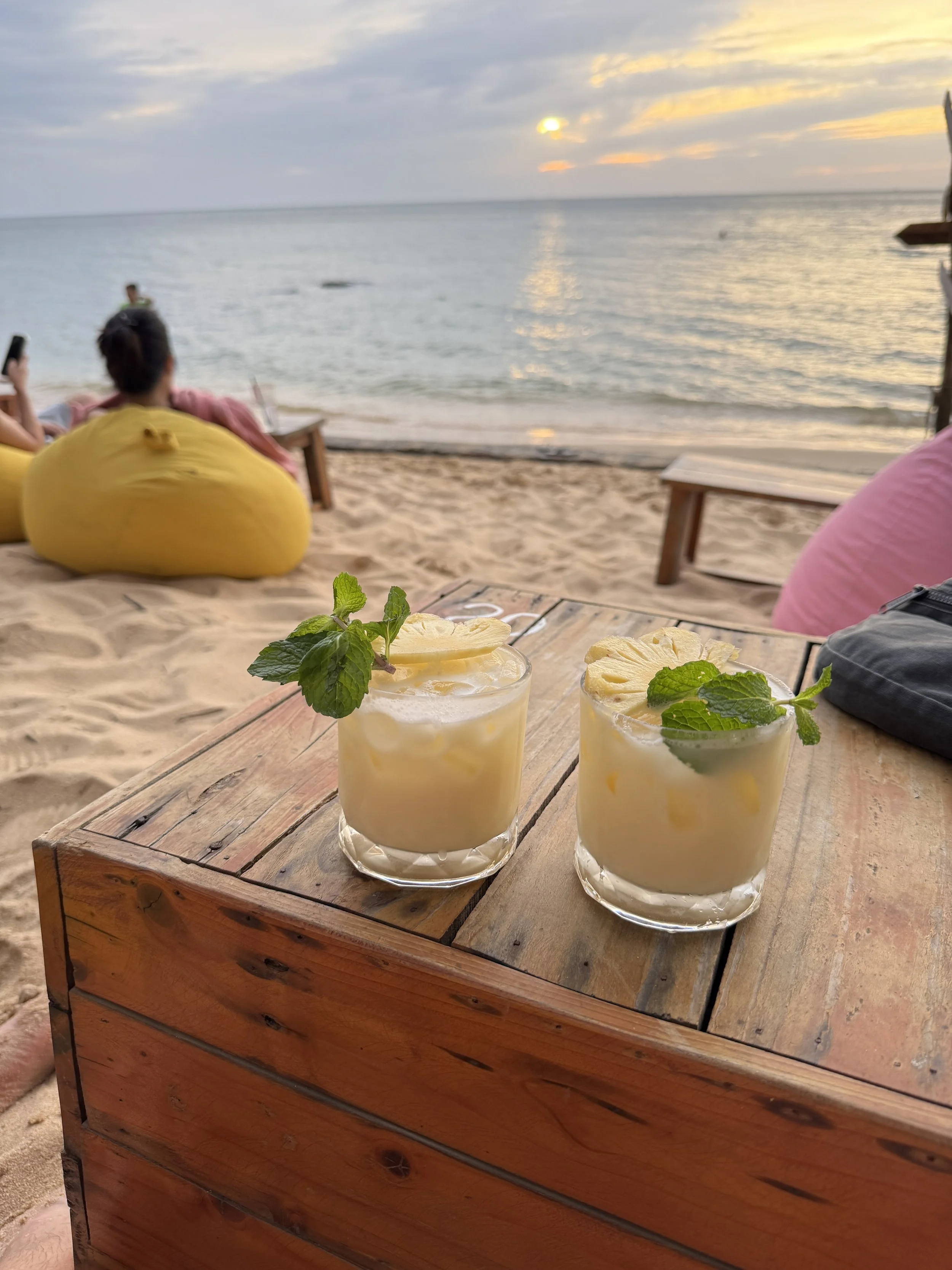 Two cocktails with lemon slices and mint garnish on a wooden table, set on a sandy beach with a sea view at sunset.