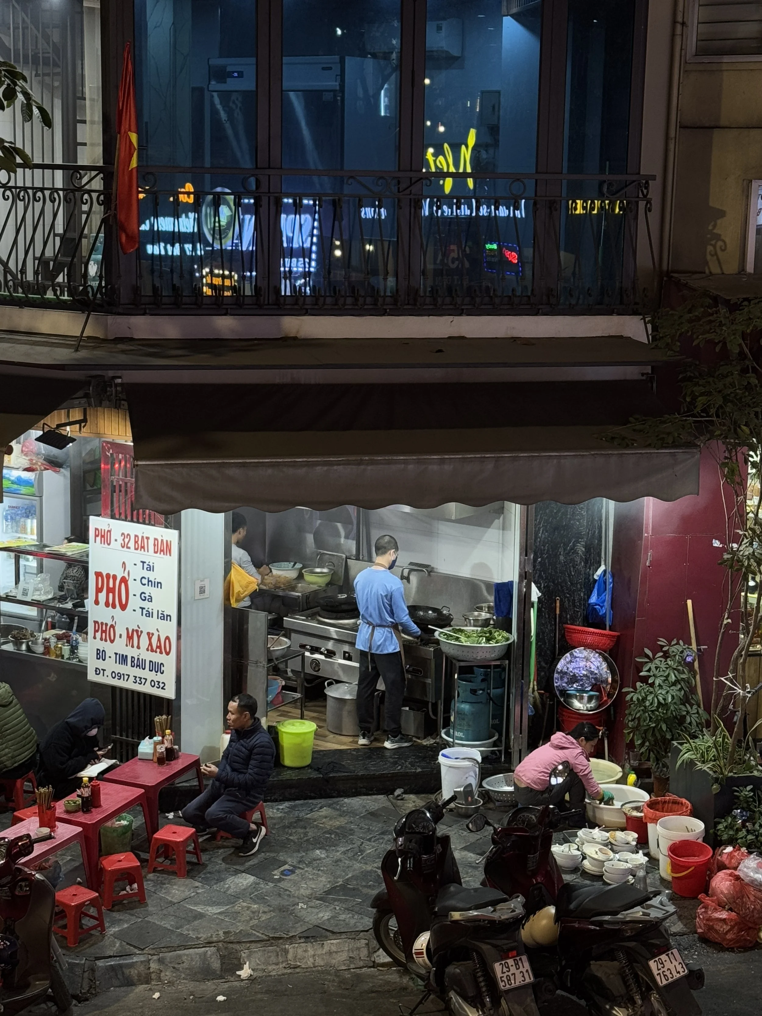 Street food stall in a Vietnamese city serving pho, with customers seated at small red tables and a cook preparing food. Motorbikes are parked nearby, and a Vietnamese flag is visible.