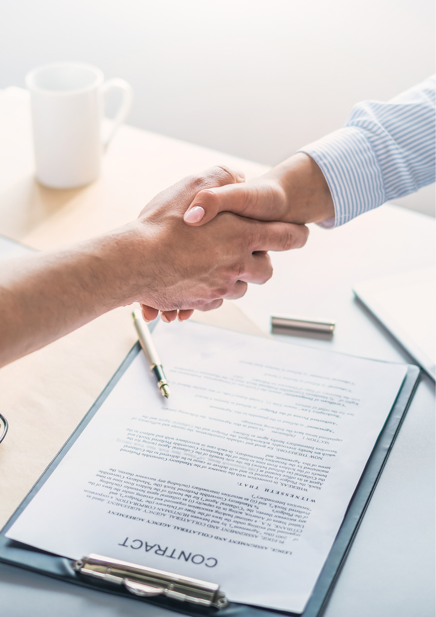 Two people shaking hands over a contract on a clipboard with a pen, with a white mug in the background.