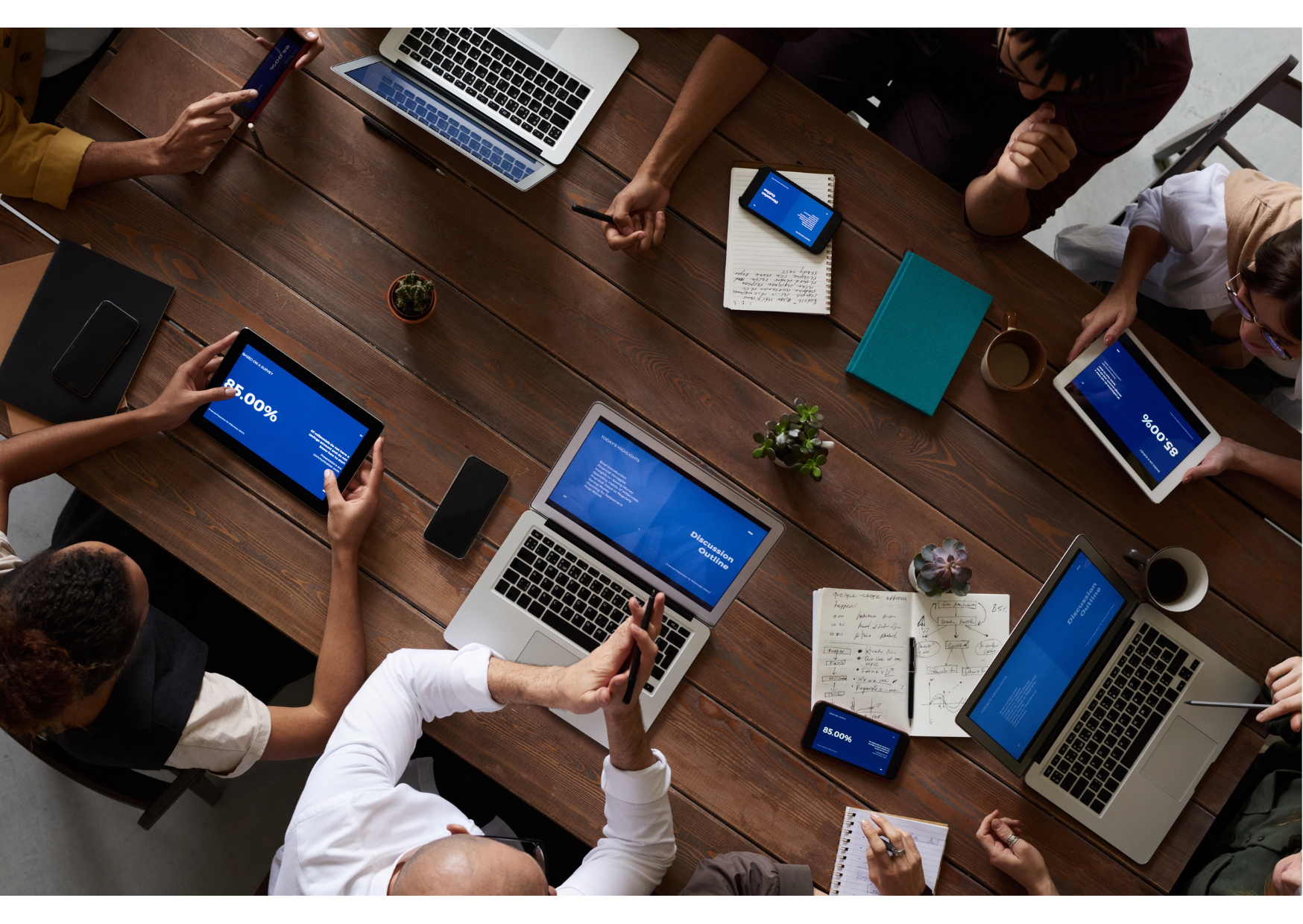 A top-down view of a business meeting with multiple people around a large wooden table, using laptops, tablets, and smartphones, with notes, notebooks, and potted plants on the table.