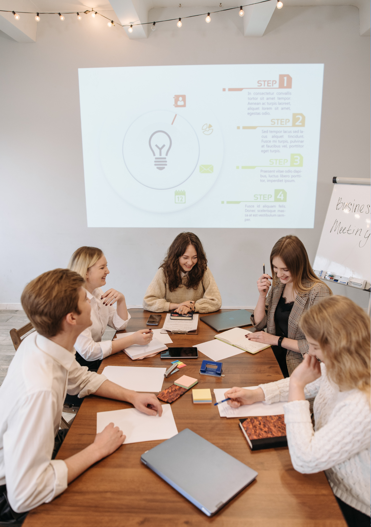 A group of six women sitting around a wooden conference table, engaged in a business meeting, with notebooks, papers, laptops, and stationery on the table. In the background, a projected presentation slide with a diagram and four steps is visible on the wall. One woman appears to be speaking, while others are smiling and listening.