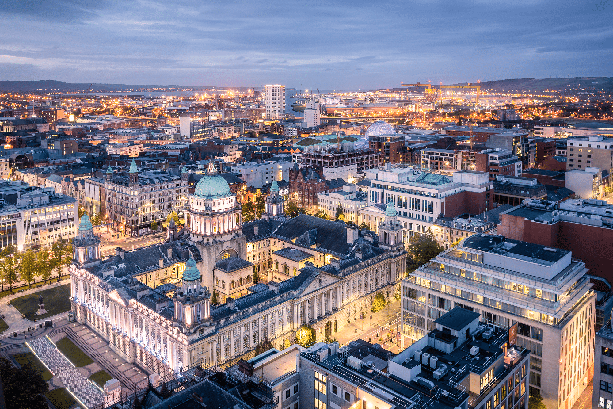Cityscape at dusk showing historic and modern buildings, with lights illuminating the scene, a river in the distance, and hills on the horizon.