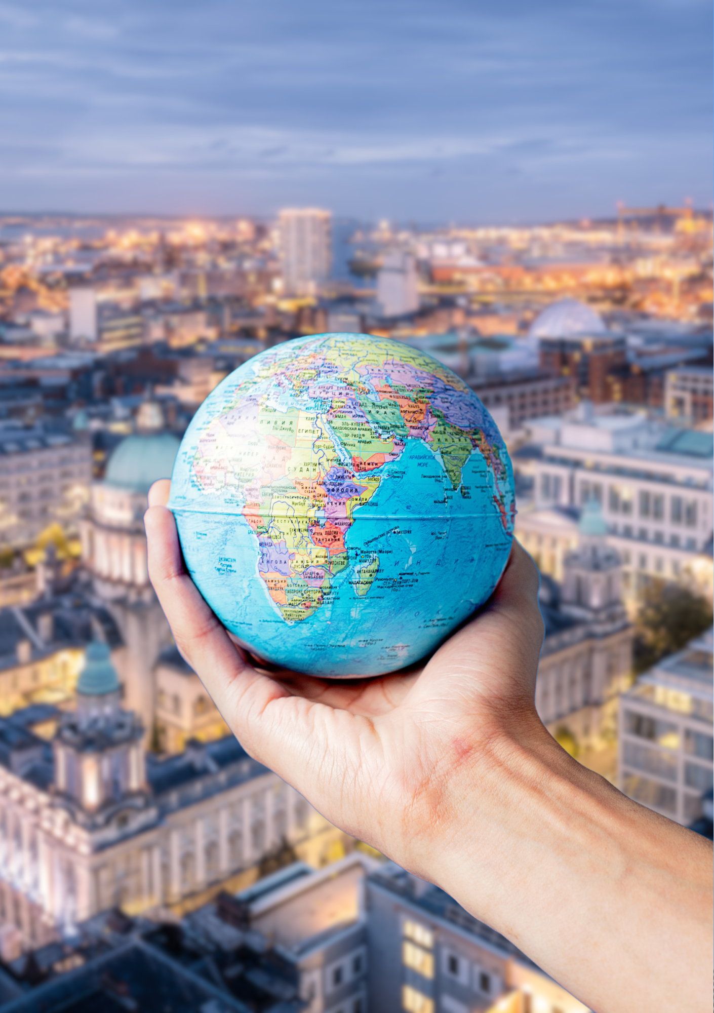 Person holding a small globe of Earth with continents visible, overlooking a cityscape with buildings and rooftops.
