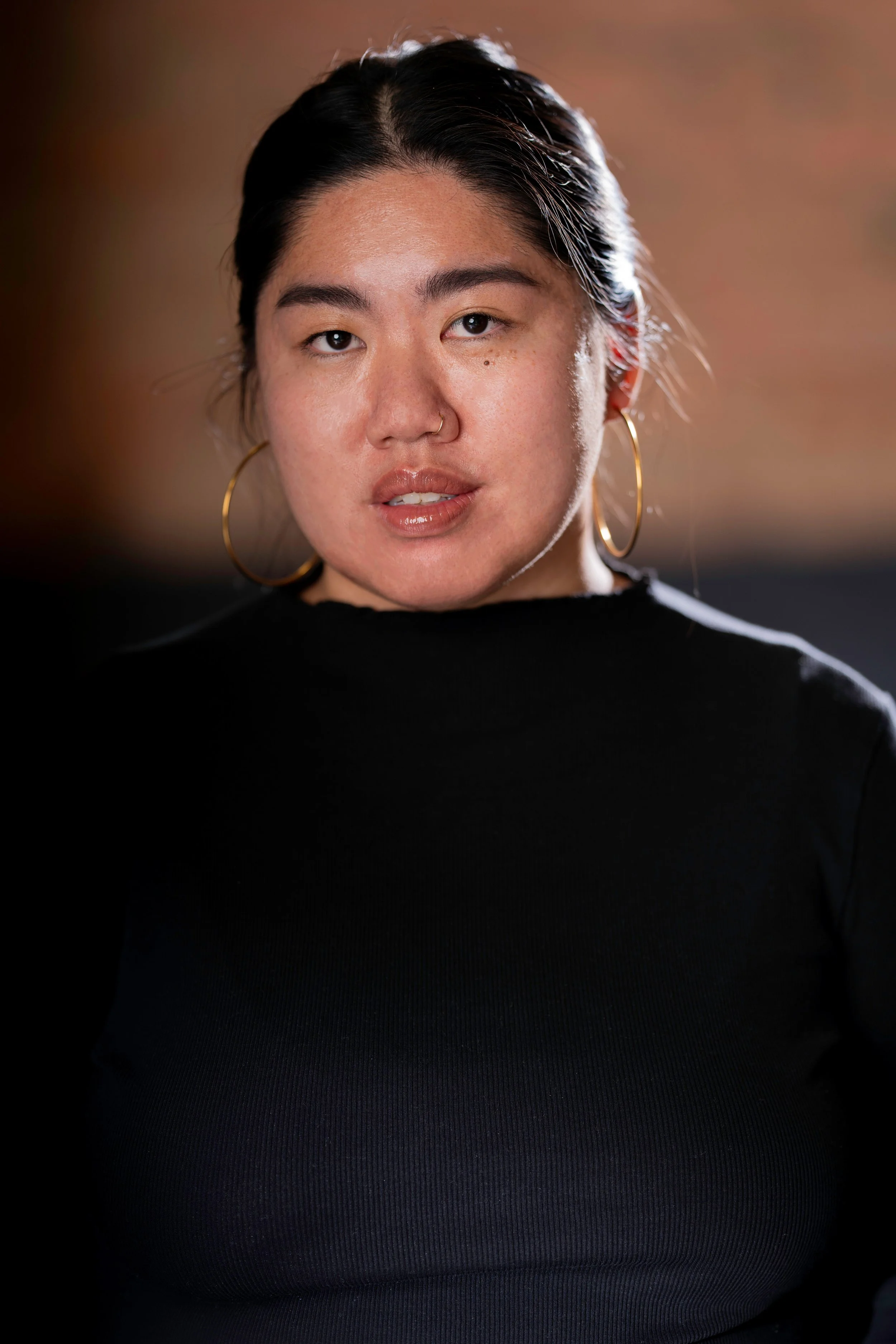 Close-up of a woman with dark hair tied back, wearing large hoop earrings, a nose ring, and a black top. She has a neutral expression and is looking directly at the camera, with a blurred warm-toned background.