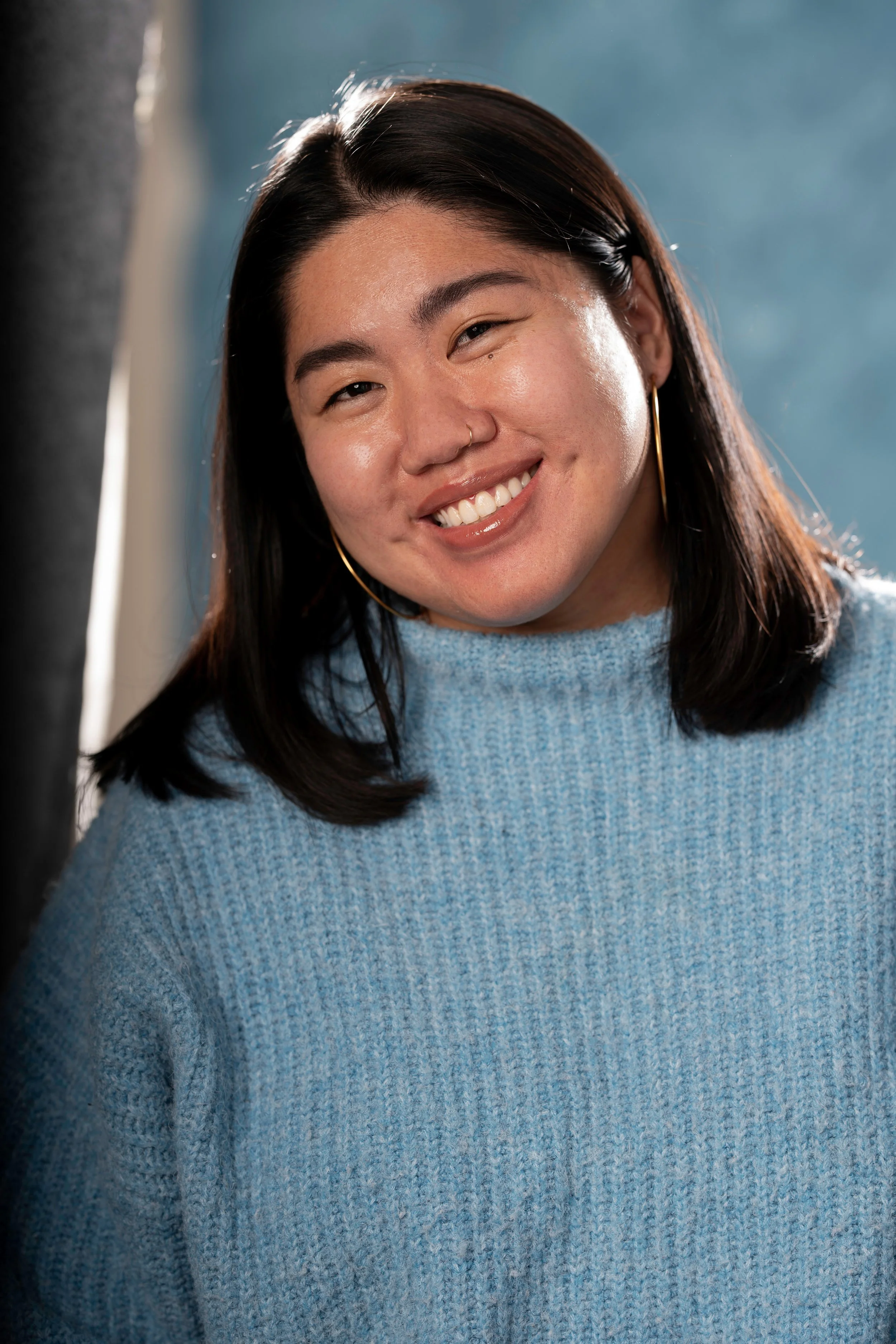 A young woman with dark hair and a nose ring, smiling and wearing a light blue knitted sweater and large hoop earrings, standing outdoors with a blue sky background.