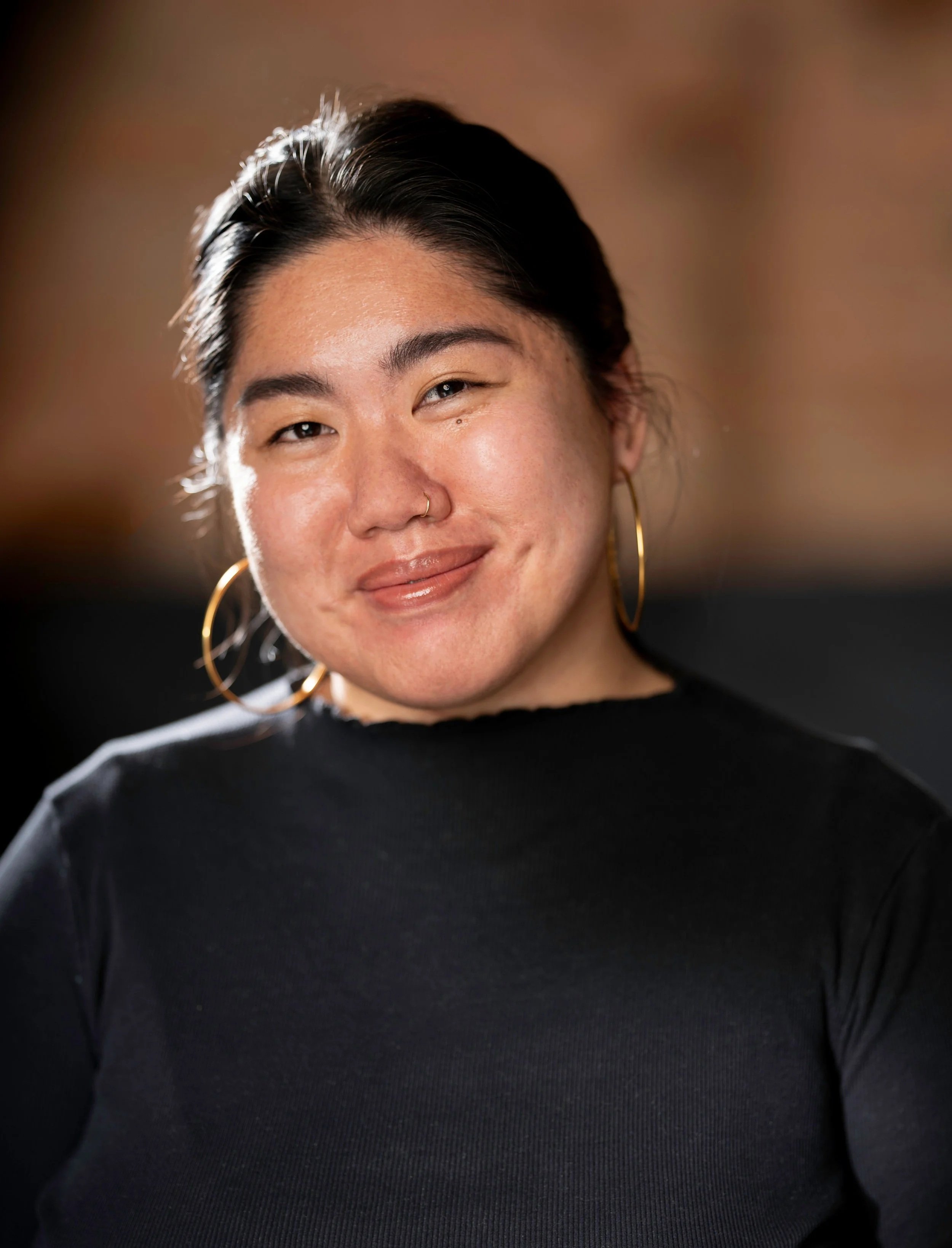 A smiling woman with dark hair, wearing gold hoop earrings and a black top, posing against a warm, blurred background.