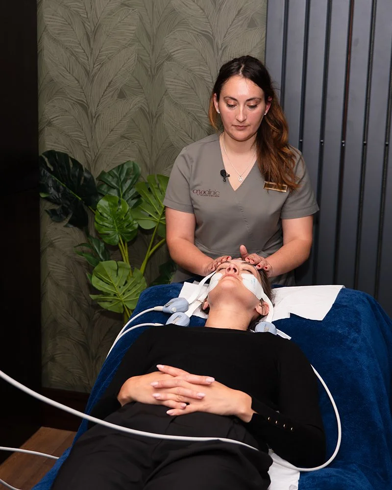 A woman with long brown hair lying on a treatment table, receiving a facial massage or treatment from a professional skincare specialist in a clinic setting with tropical leaf wallpaper and green plants in the background.