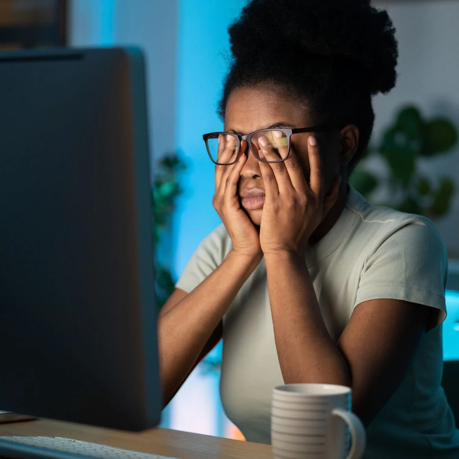 Frustrated woman wearing glasses, holding her face with both hands while sitting at a desk in front of a computer.