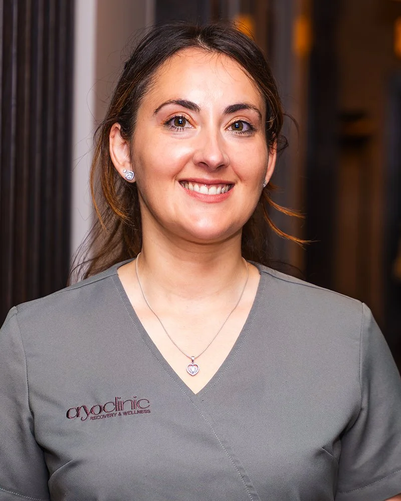 A smiling woman with brown hair, wearing earrings and a necklace, dressed in gray scrubs