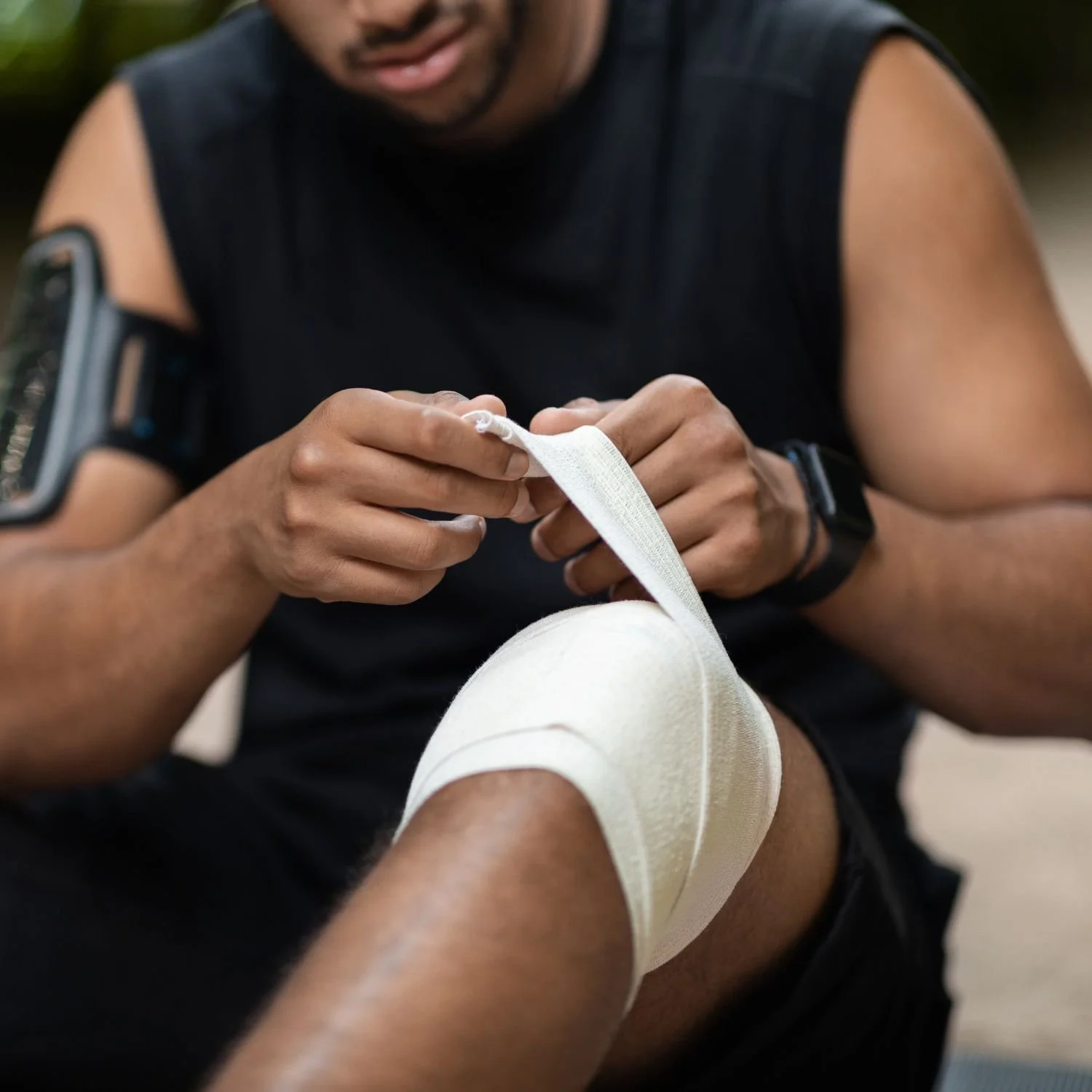 A man wearing a black sleeveless shirt and a smartwatch is wrapping a white athletic tape around his knee.
