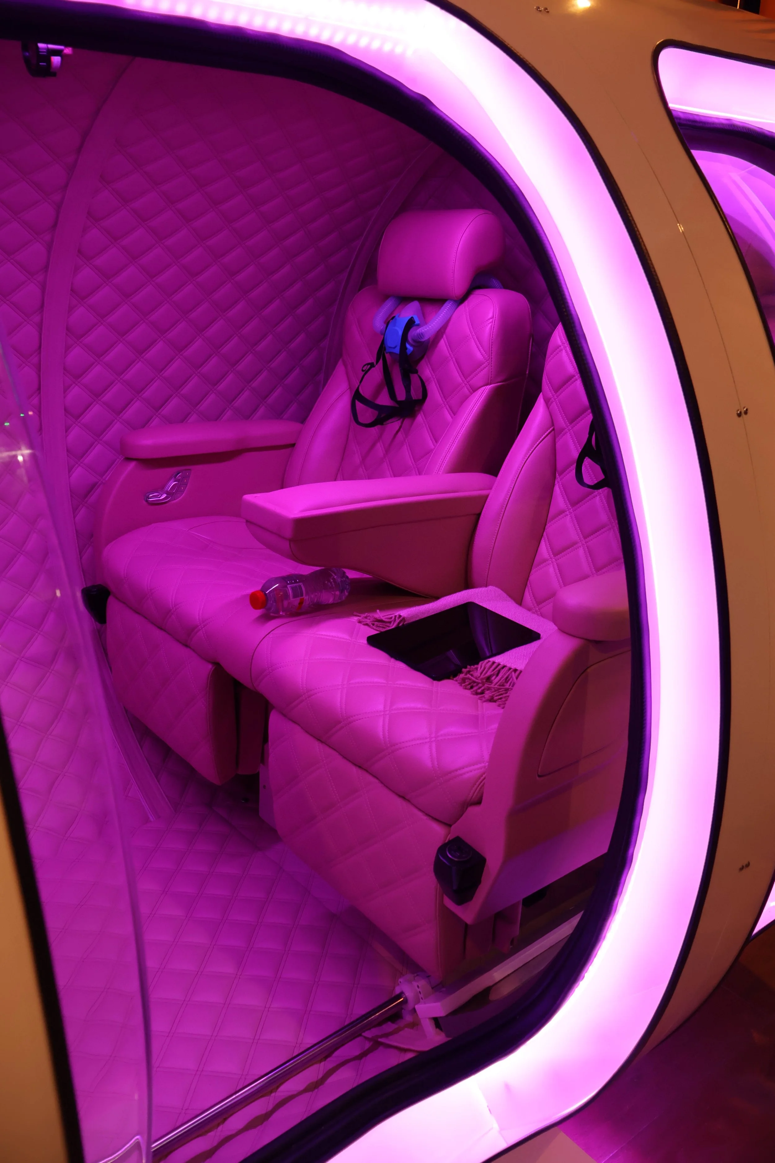 Interior of a capsule with pink quilted seats and walls, illuminated by pink and purple LED lights, featuring headphones hanging on the headrest and a water bottle on the seat.
