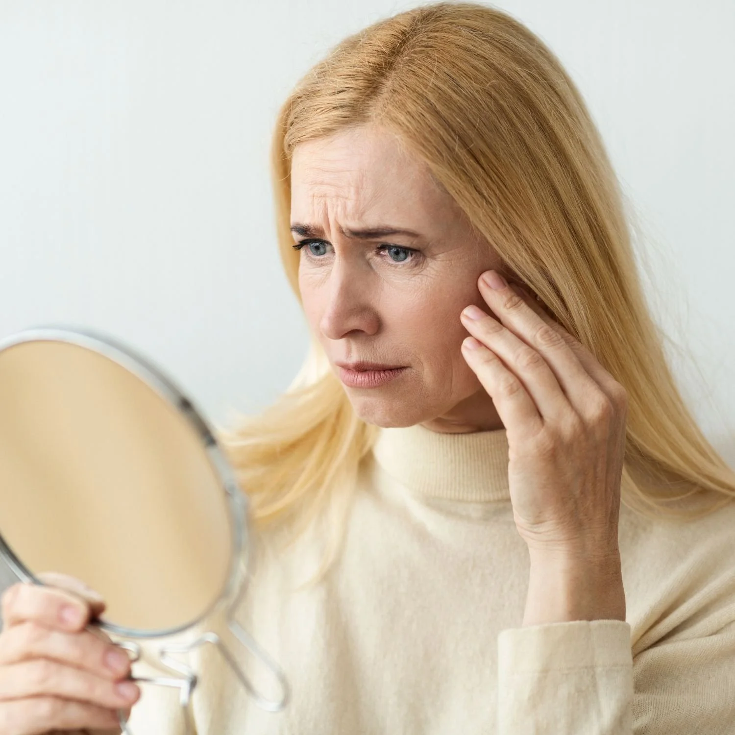 A woman with long blonde hair looking concerned while holding her cheek and examining her face in a mirror.