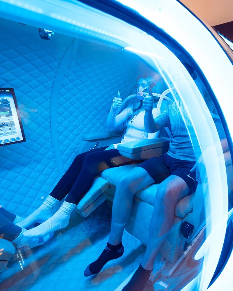 Two adults inside a HBOT chamber at CryoClinic, surrounded by blue lights, in a wellness facility.