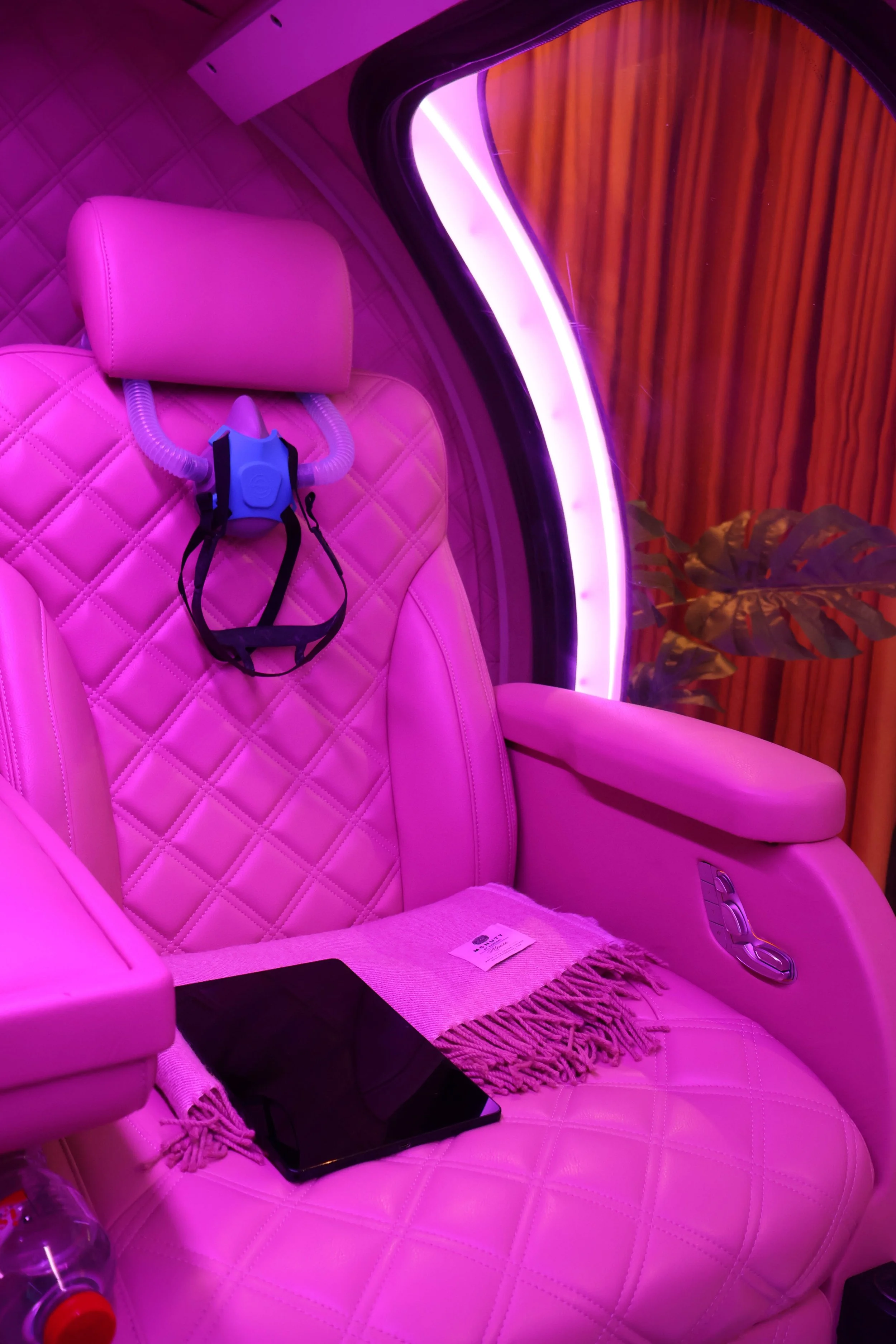 A pink quilted recliner chair with a headrest, a mask hanging on the side, a tablet, a piece of cloth, and a bottle on the armrest. The chair is surrounded by pink and purple lighting, with a background of wooden curtains and a potted plant.