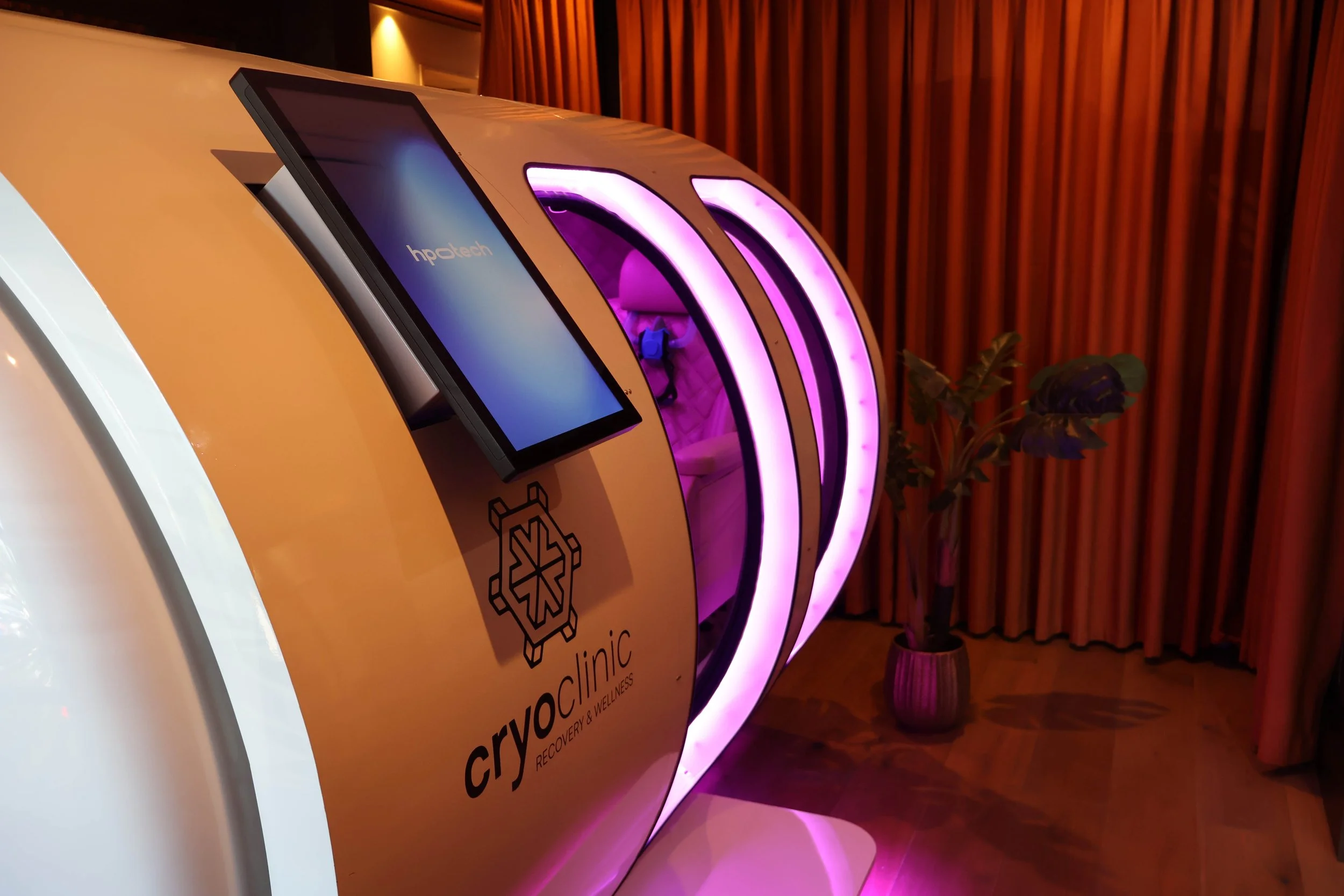 A promotional display features a flourescent lit glowing purple ring next to a monitor. The display also has a logo of a snowflake with a heartbeat line inside and the words 'Cryo Clinic' and 'Recovery & Wellness' on it. The setting appears to be ind