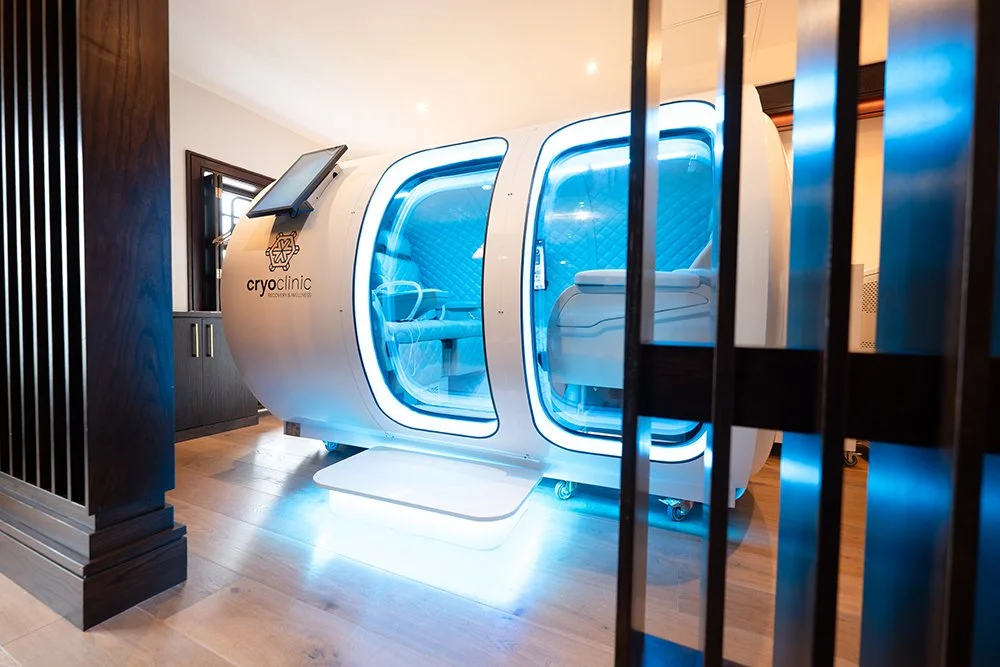 A modern HBOT chamber with blue lighting, labeled 'CryoClinic.' It is in a room with wooden floors and dark wood paneling.