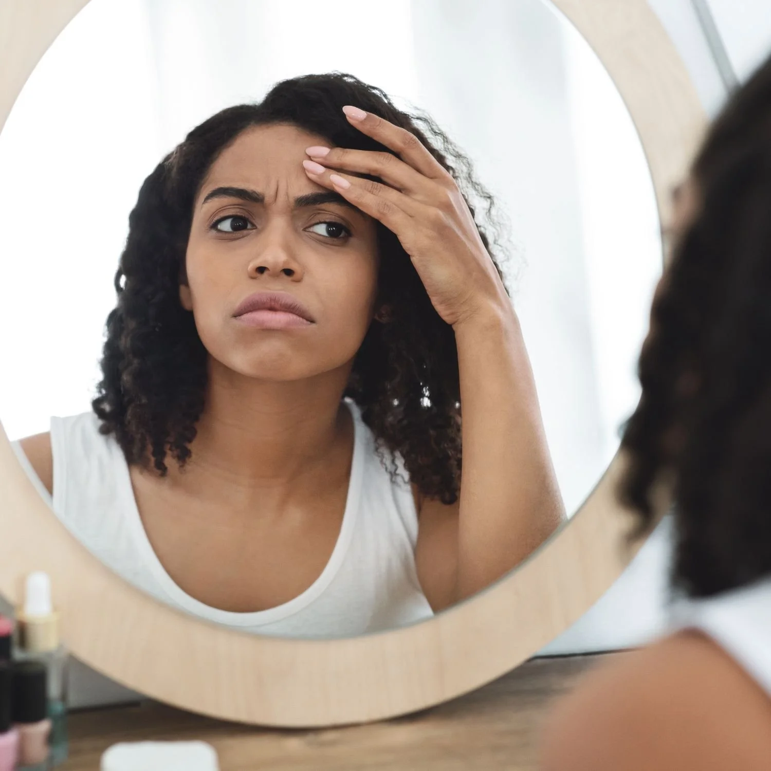 Young woman with curly hair looks at her reflection in a mirror, feeling worried or stressed, with her hand on her forehead.