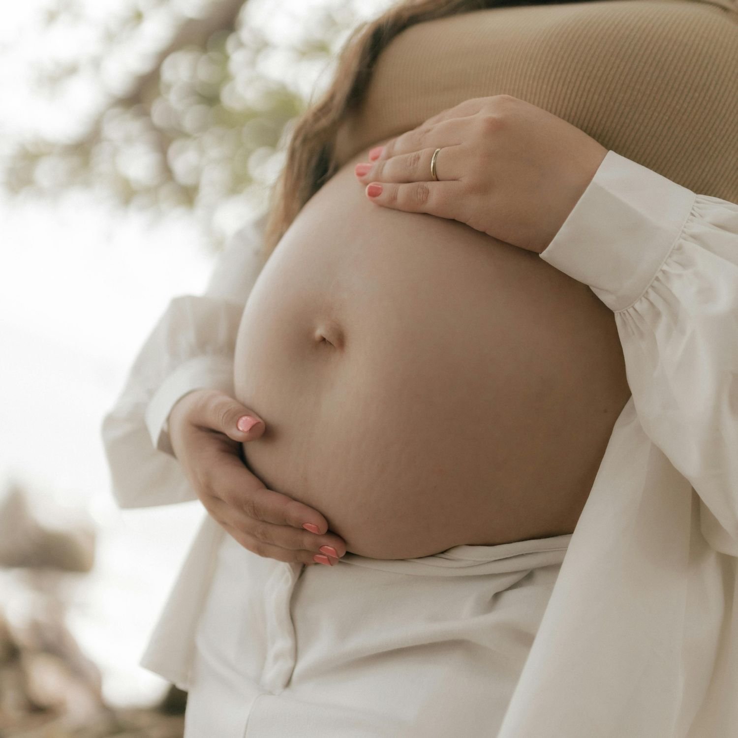 Close-up of a pregnant woman touching her belly outdoors, wearing a white shirt and a beige sleeveless jacket.