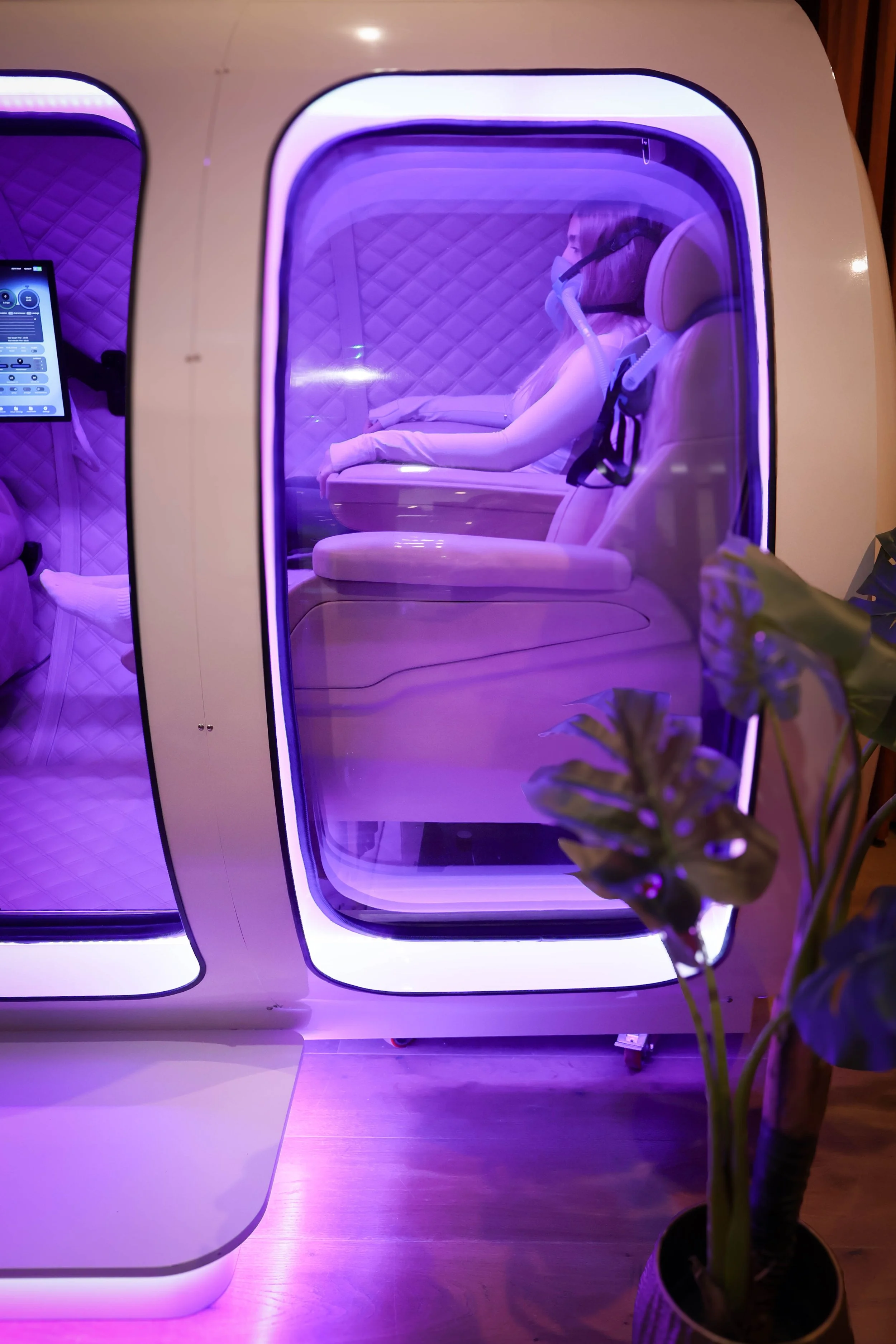 A person sitting Inside an HBOT capsule with purple lighting, wearing a mask and lying on a cushioned surface
