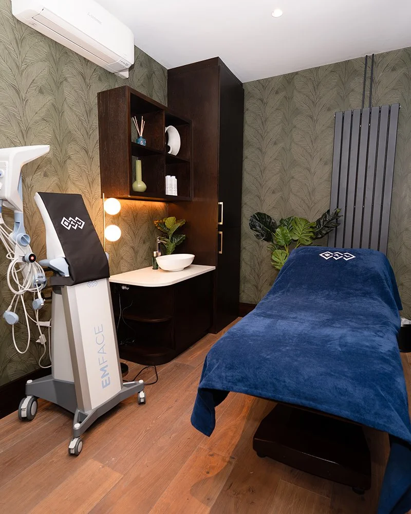 A wellness or spa treatment room with a massage bed covered in a blue towel, a facial or treatment machine labeled 'EMFACE', a dark wood cabinet with plants and decorative items, a wall-mounted air conditioner, and wallpaper with a leafy pattern.