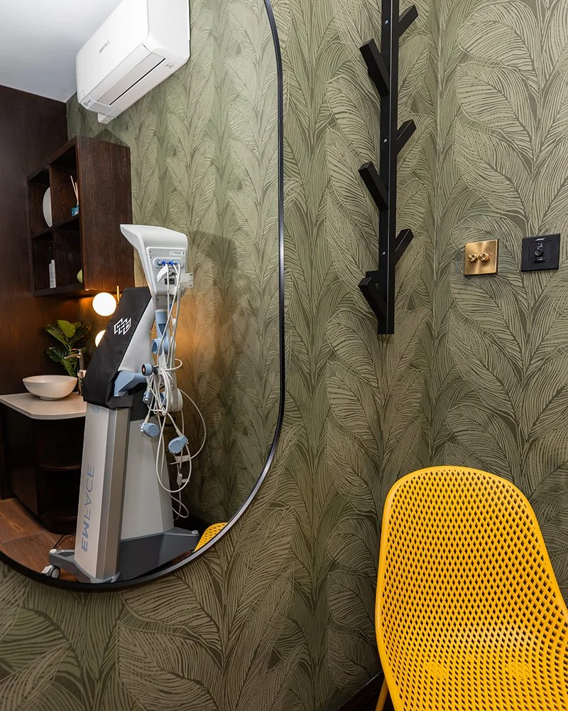 A round mirror reflecting a room with a medical or dental chair at an angle. The room has leafy wallpaper, a wooden shelf with decorations, a bowl on a small table, and a yellow chair. There is an air conditioning unit on the wall, and various electr
