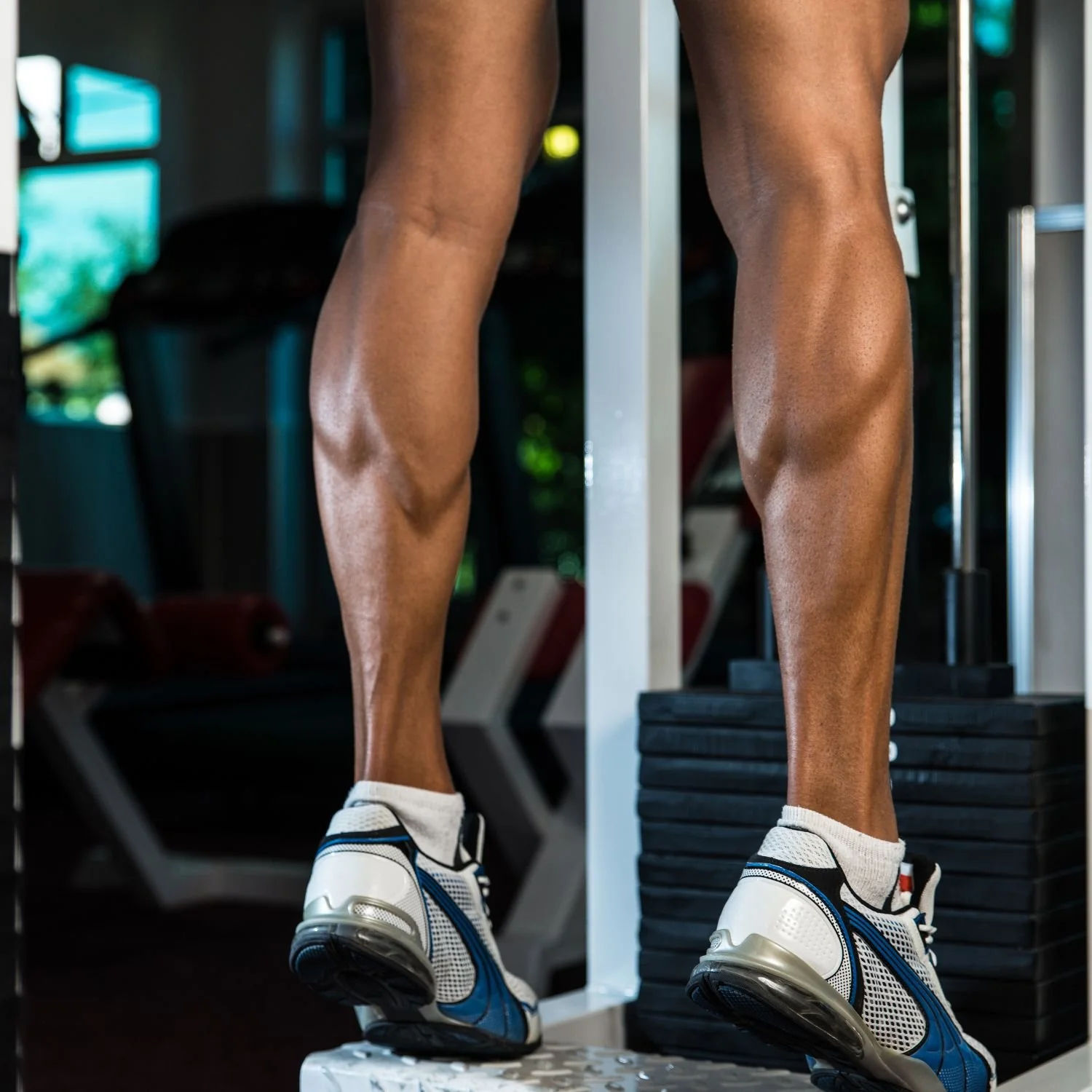 Close-up of a person's muscular legs in athletic shorts, standing on the edge of a treadmill in a gym, with exercise equipment in the background.