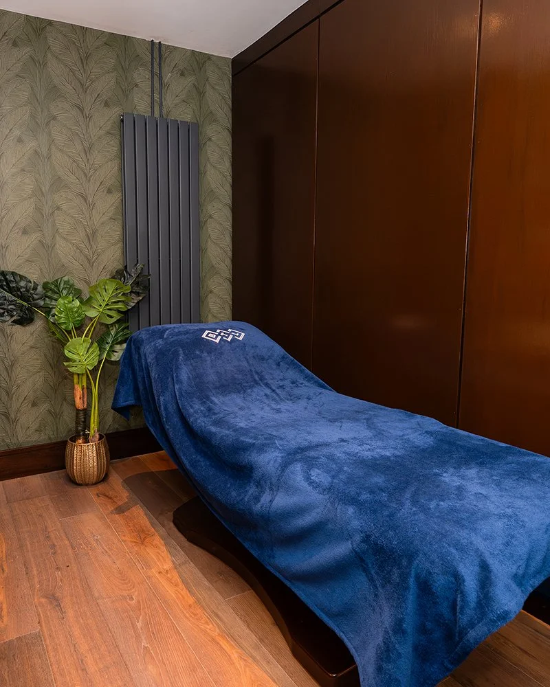 Treatment bed covered with a blue blanket in a room with wooden flooring, green leafy wallpaper, a modern black radiator, and a large wooden closet or wall panel.