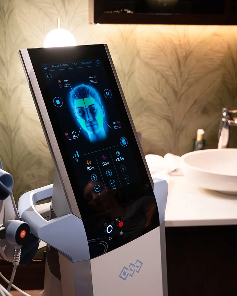 An EMFace device with a digital screen displaying a diagram of a human face, showing settings for therapy. The device is in a room with a patterned wall, a table with a bowl-shaped sink, and a lamp.